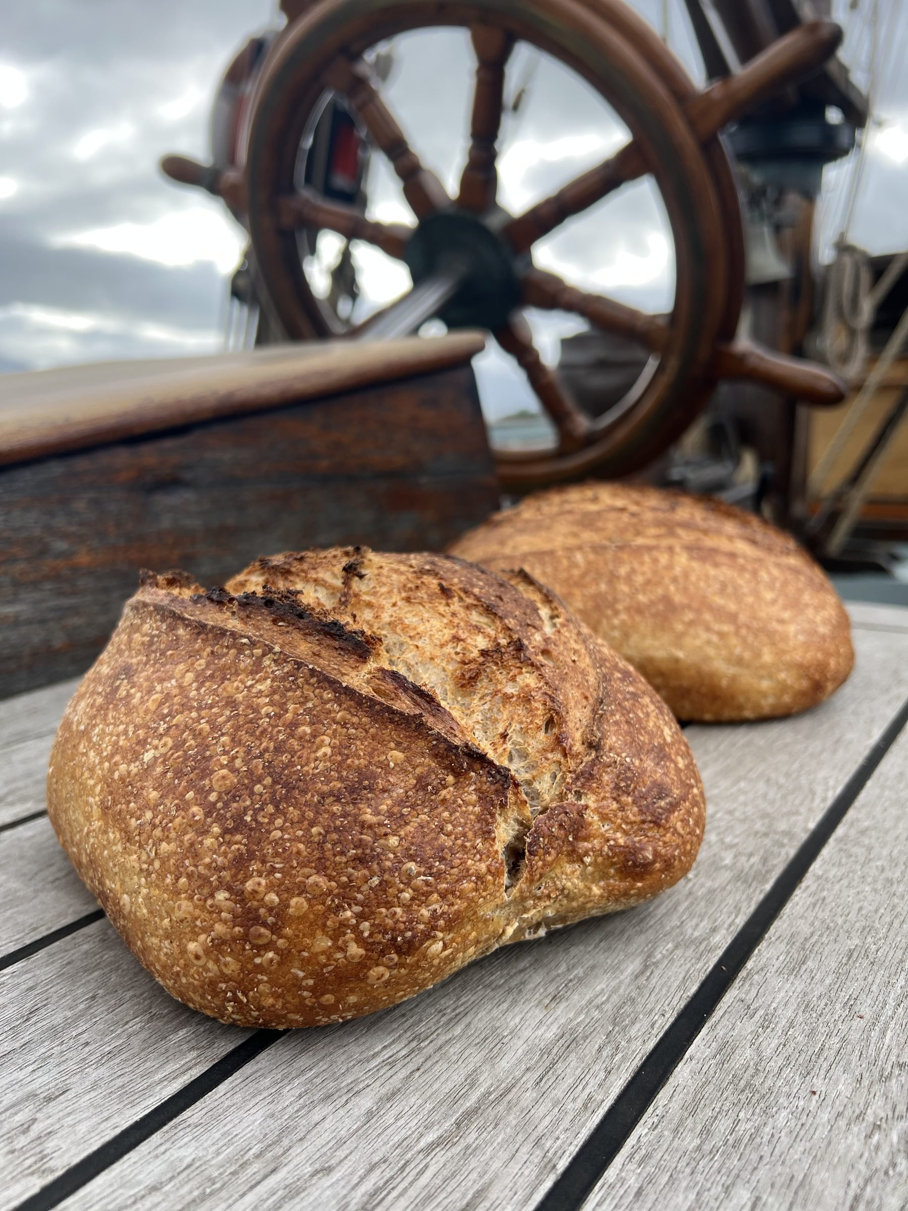 Close-up of a loaf of bread on a wooden deck, with a wheel of a ship's helm in the background.