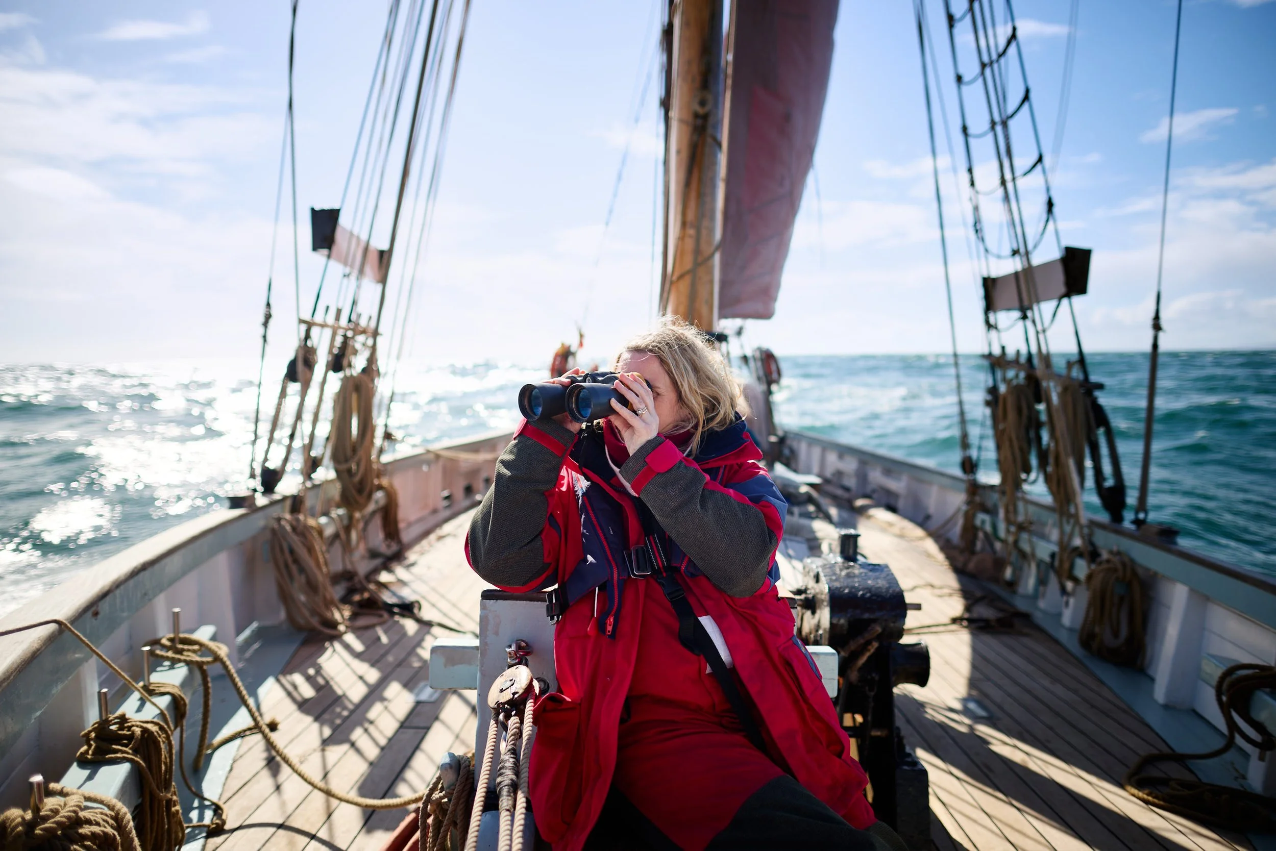 Woman in red jacket and glasses holding binoculars while on a sailing ship, with rigging and the ocean visible.