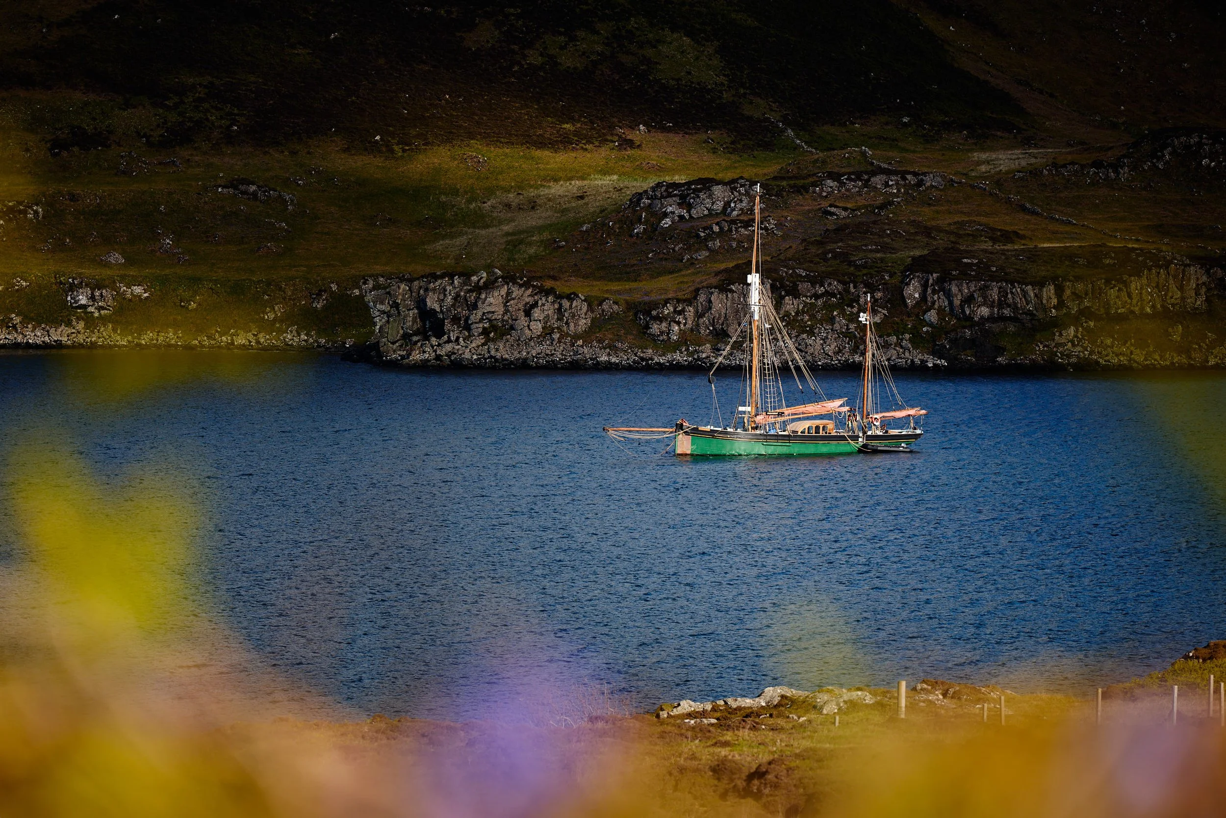 A sailboat with a green hull on a calm body of water, against a backdrop of rocky cliffs and hillside covered in green and brown vegetation.
