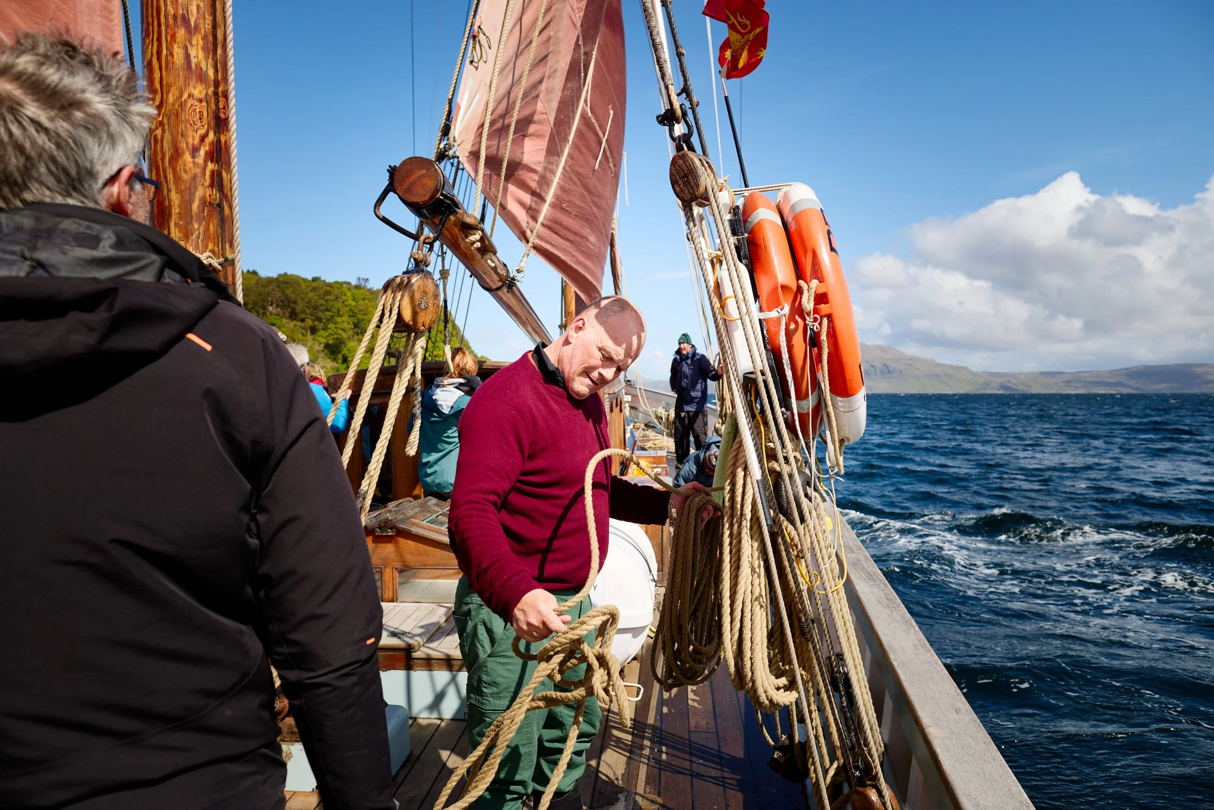 People on a boat, with one man in a red sweater handling ropes, and others in the background, sailing on a body of water under a partly cloudy sky.