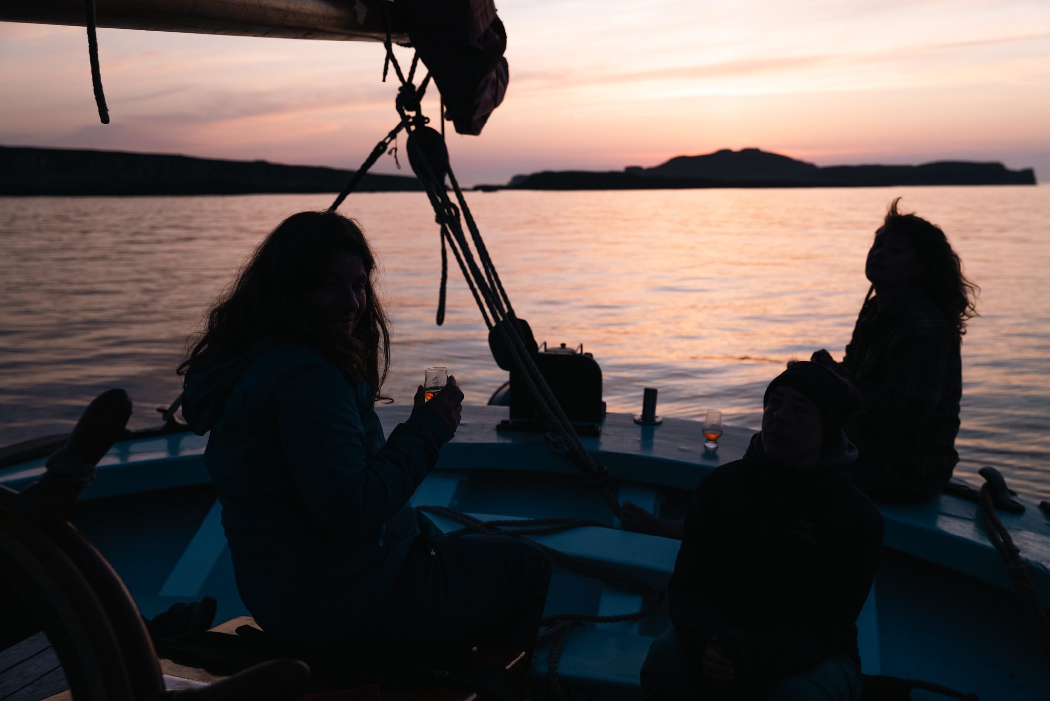People enjoying a sunset on a boat, with a woman holding a glass of wine and others sitting around, silhouetted against the colorful sky and calm water.