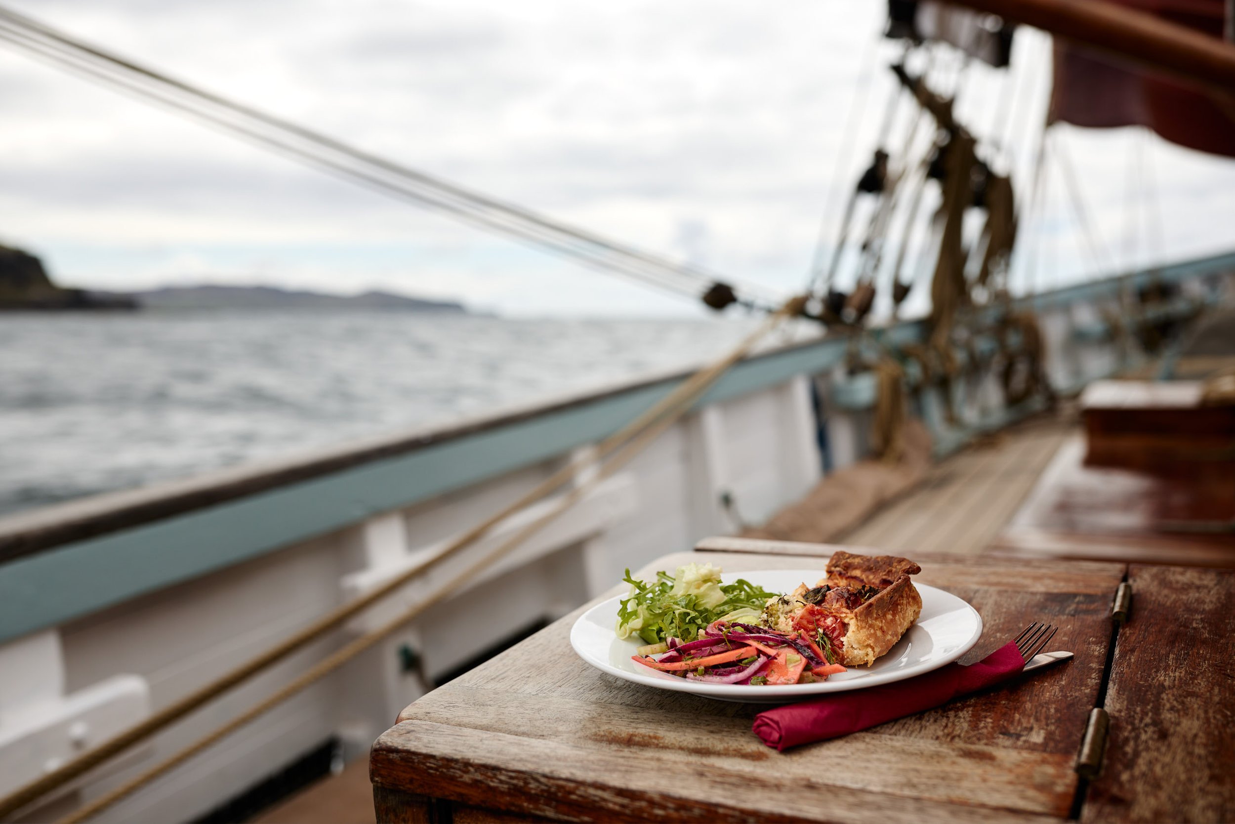 A white plate with a slice of vegetable quiche and a side salad with greens, tomatoes, and purple onions, placed on a wooden table outdoors with a view of water and clouds.