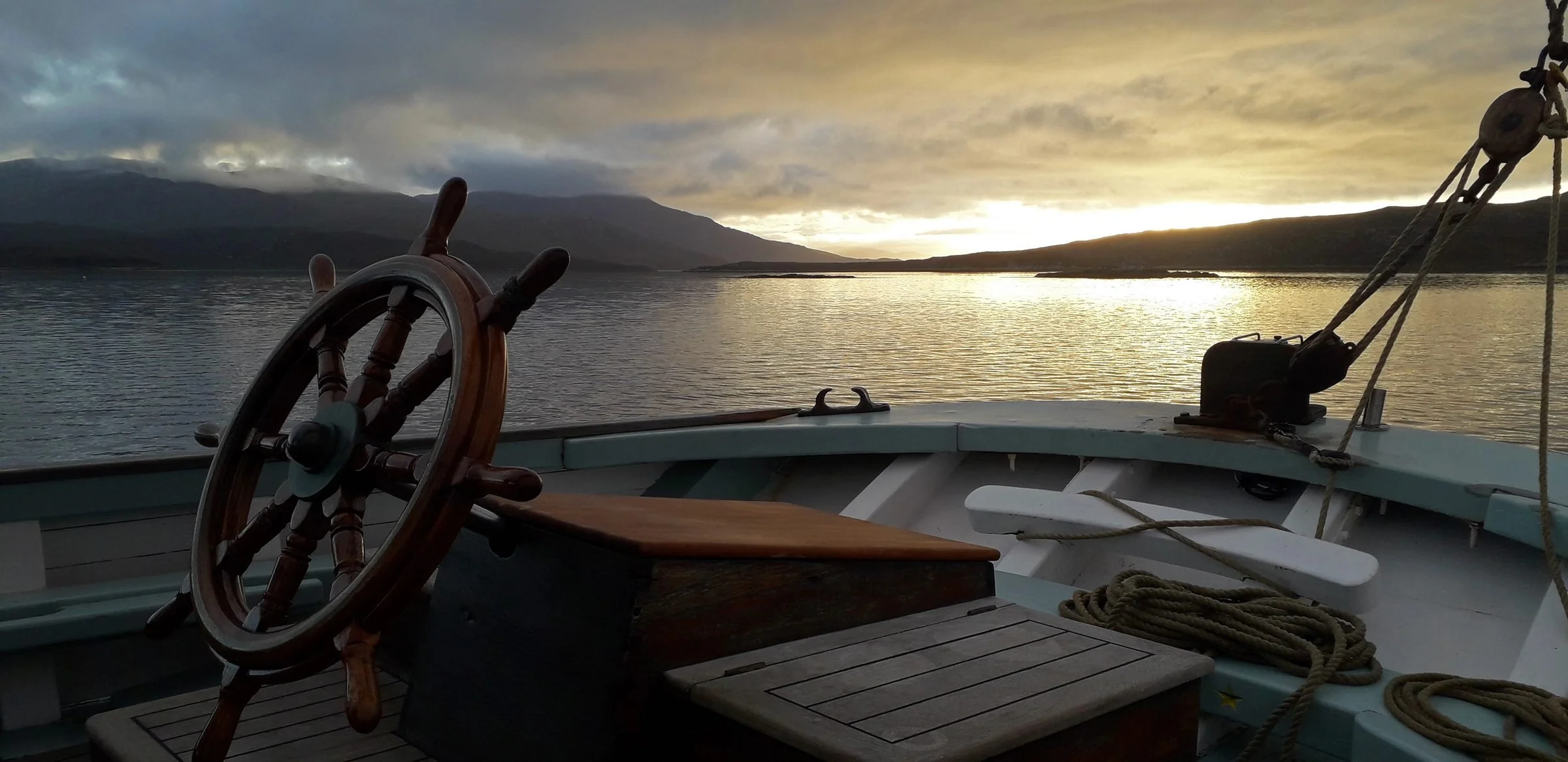 View from Provident showing a steering wheel, ropes, and wooden bench on a calm lake during sunset with mountains and cloudy sky in the background.