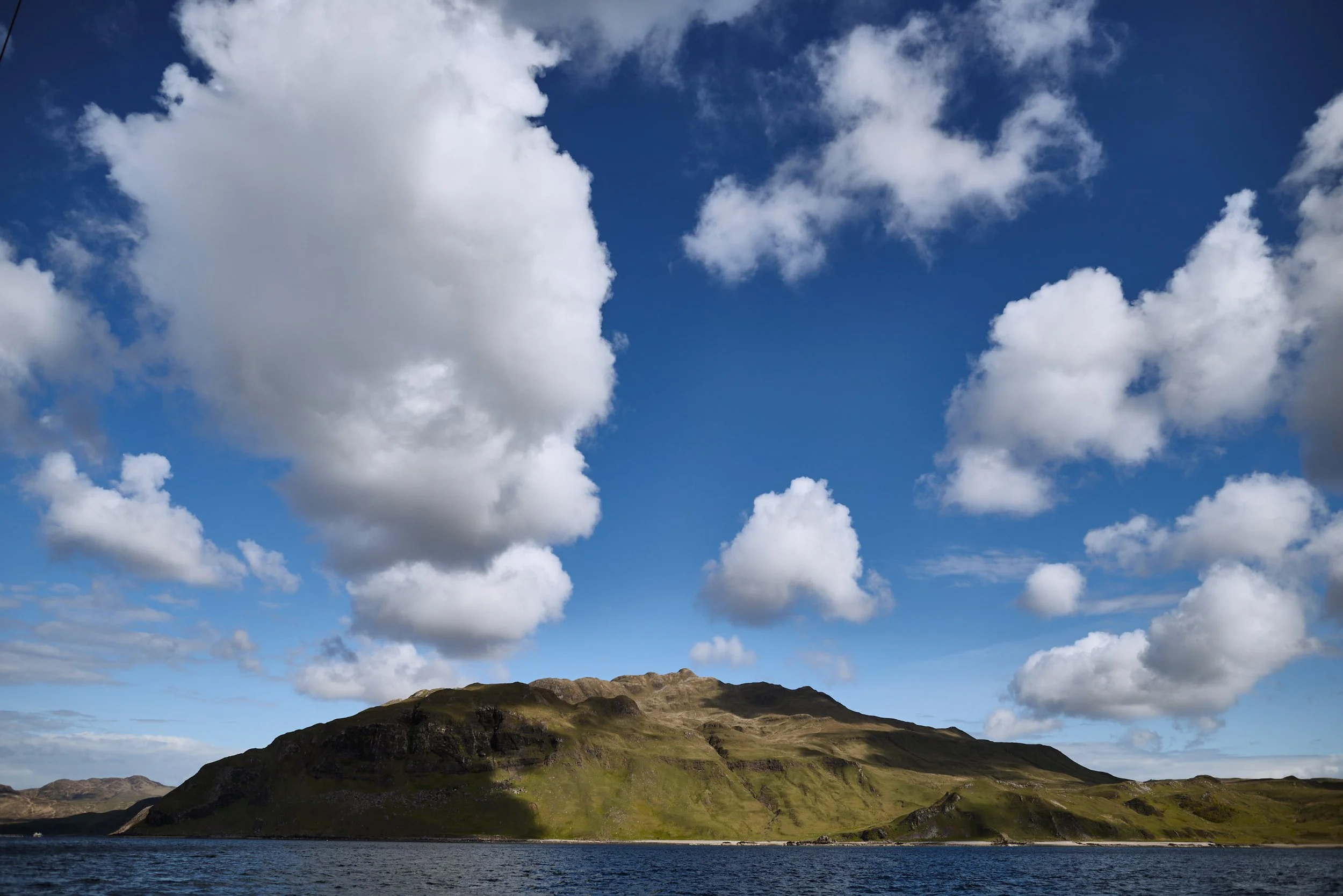 A landscape with a body of water in the foreground and a hillside covered with green vegetation and rocky outcrops under a partly cloudy sky.