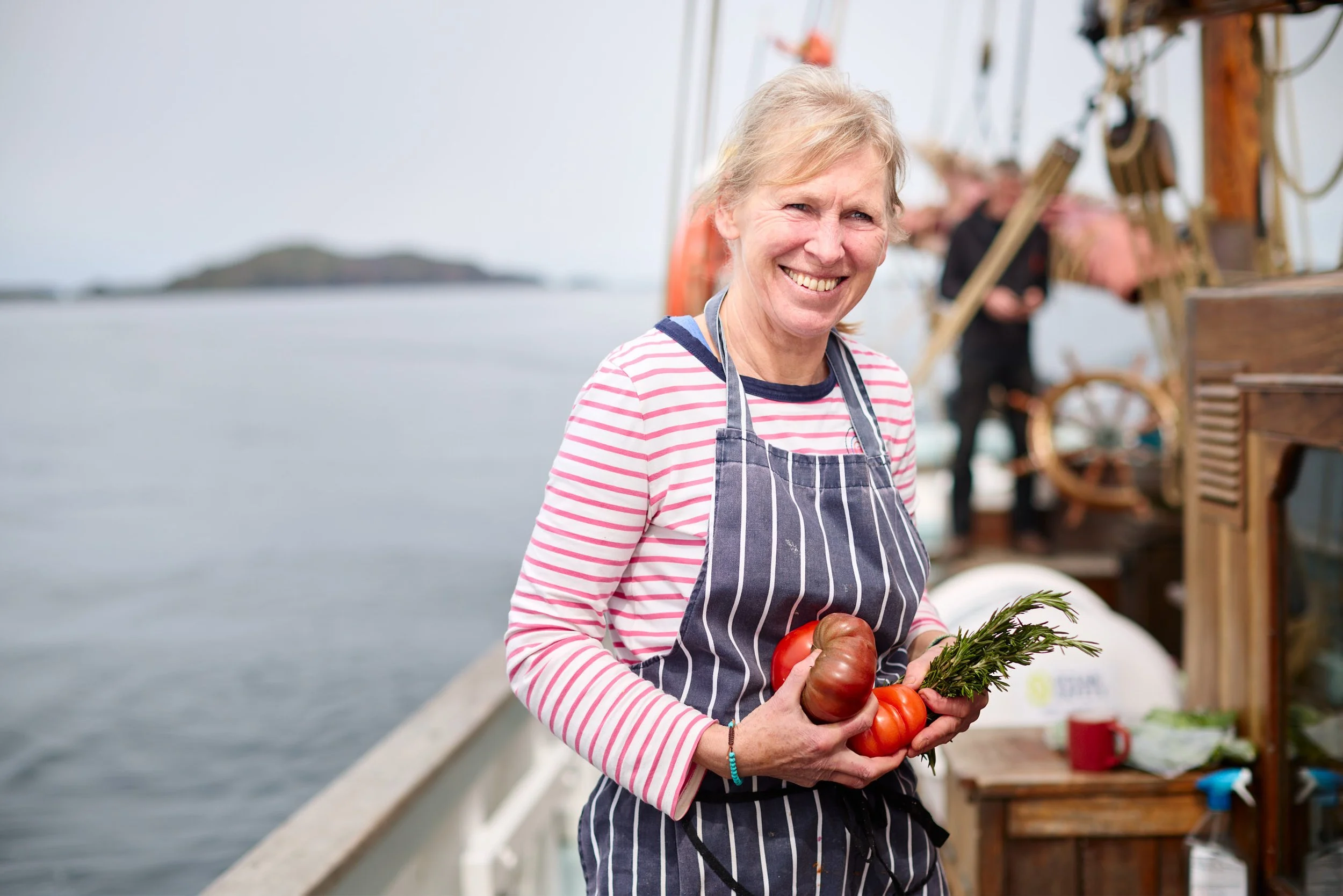 A smiling woman wearing a striped shirt and apron holding tomatoes and greenery on a boat or dock near water with an island in the background.