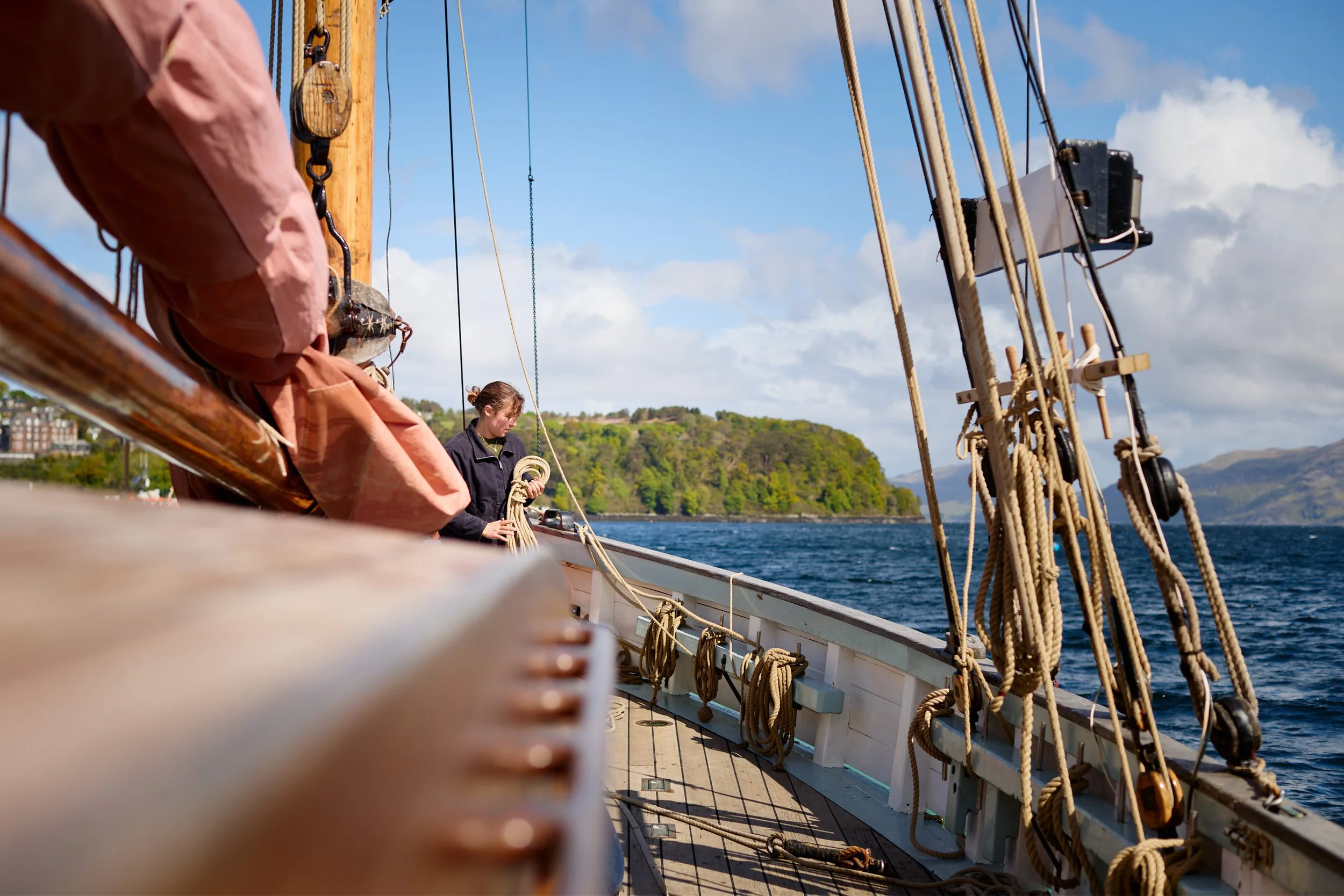 View of a sailing ship's deck with ropes, wood, and rigging, with two people working on the ship, and a scenic coastline with trees and buildings in the background under a partly cloudy sky.