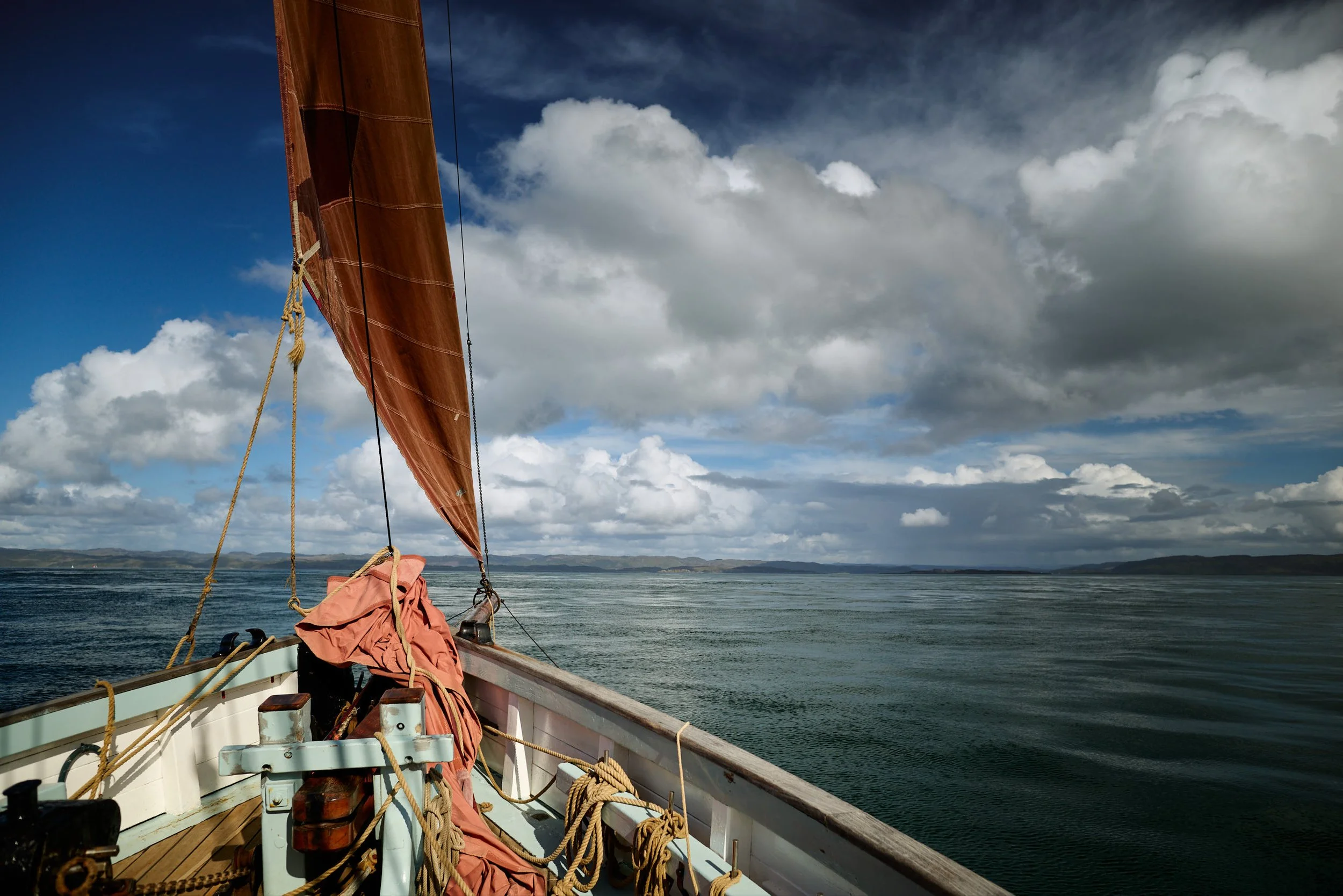 View from a sailboat's deck, provident  showing a red sail, ropes, and equipment, with calm water and a partly cloudy sky in the background.