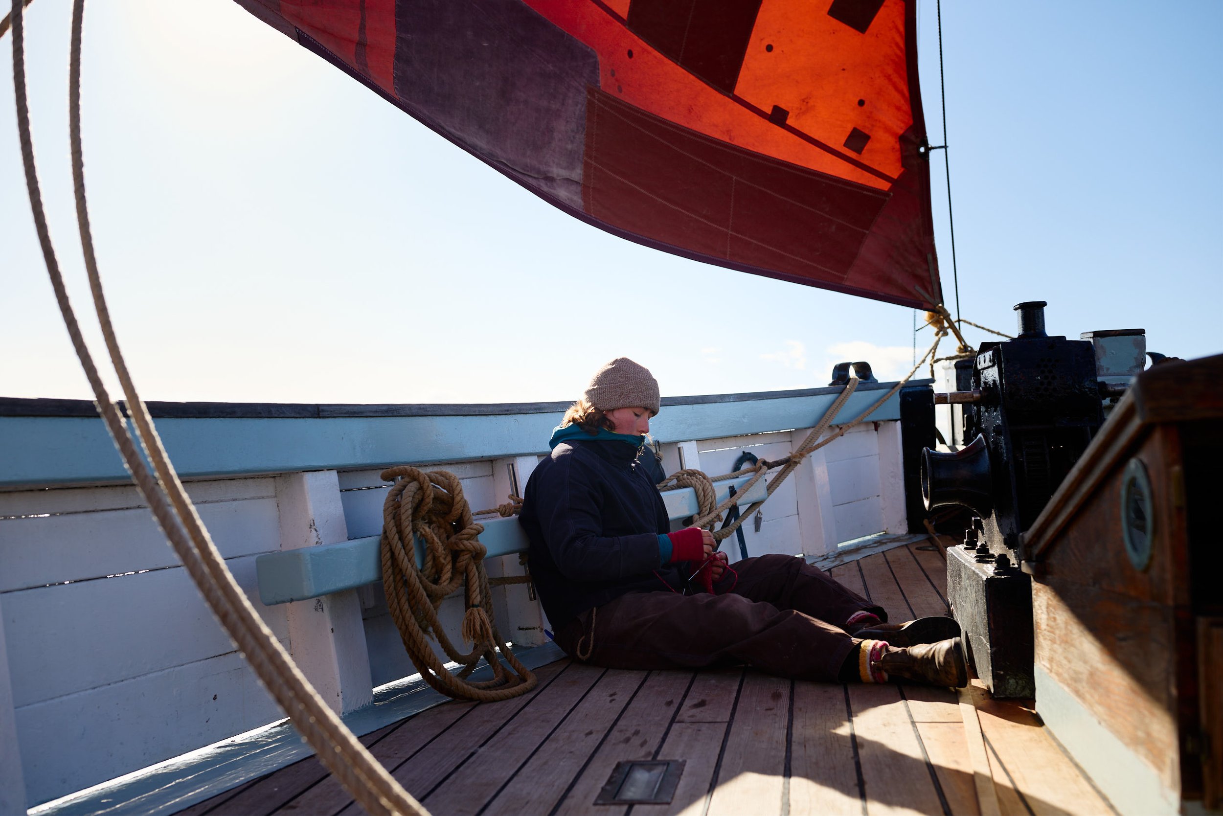 A person sitting on the deck of a boat or ship, wearing a beige knit hat, dark jacket, and red gloves, with ropes and boat equipment around them, under a large red sail, with clear blue sky in the background.