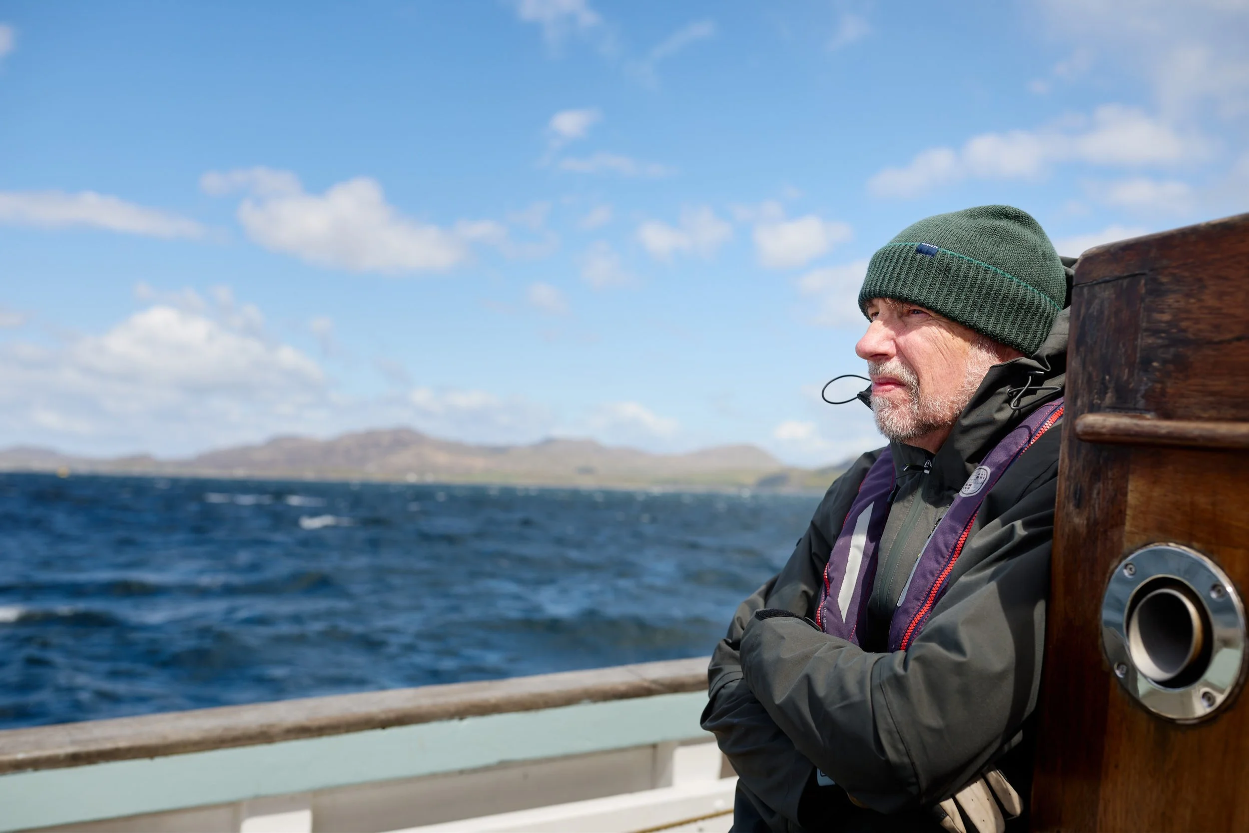 An older man with a gray beard and mustache wearing a green beanie and dark jacket sitting on a boat with a wooden railing, looking out at the water and the distant hills under a partly cloudy sky.