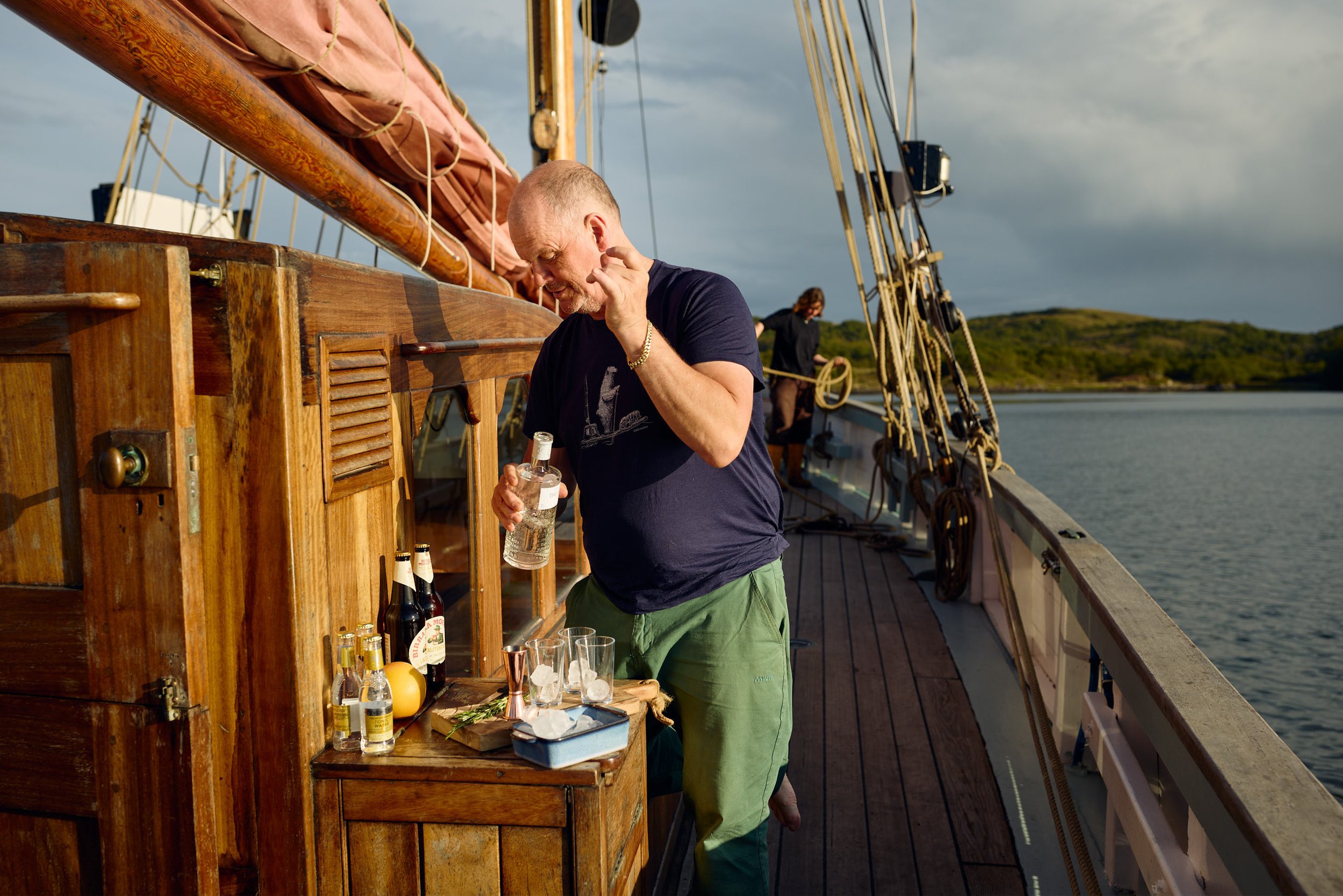 A man with short gray hair and a beard is standing on a boat near a wooden bar, holding a bottle of clear liquor and a glass with ice, while an Asian woman in black shirt is in the background handling ropes. The boat is on a body of water, with hills and a cloudy sky in the background, and the scene suggests a relaxed social setting.