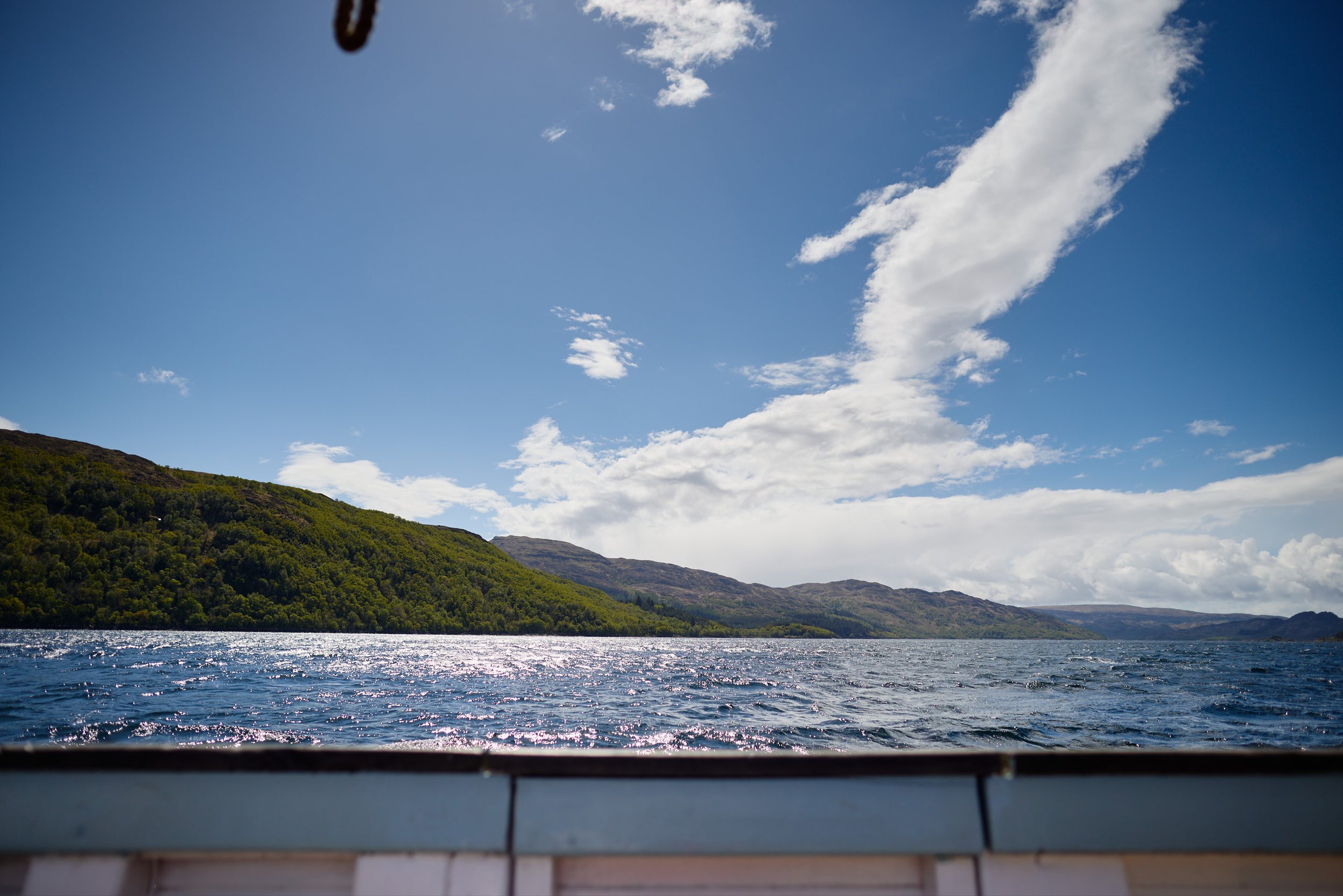 Photo taken from a boat showing a landscape with a lake, green hills, and a blue sky with scattered clouds.