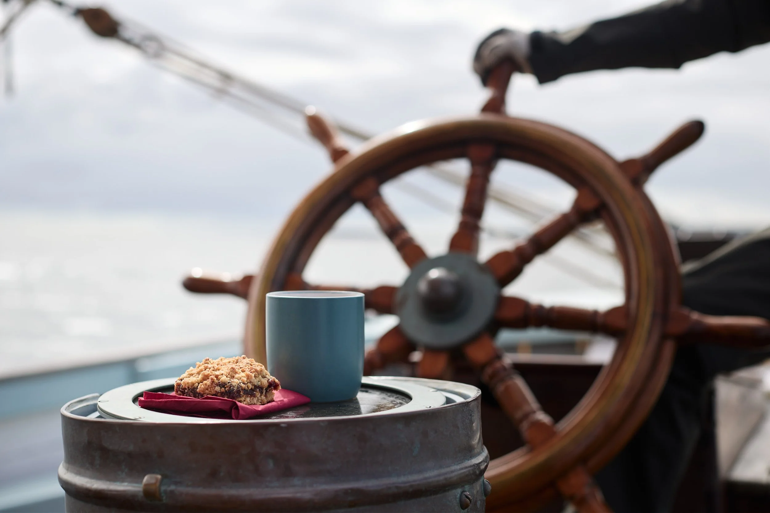 A table with a plate holding a baked dessert, a blue mug, and a napkin, set outdoors on a boat deck with a ship's wheel in the background and a cloudy sky.