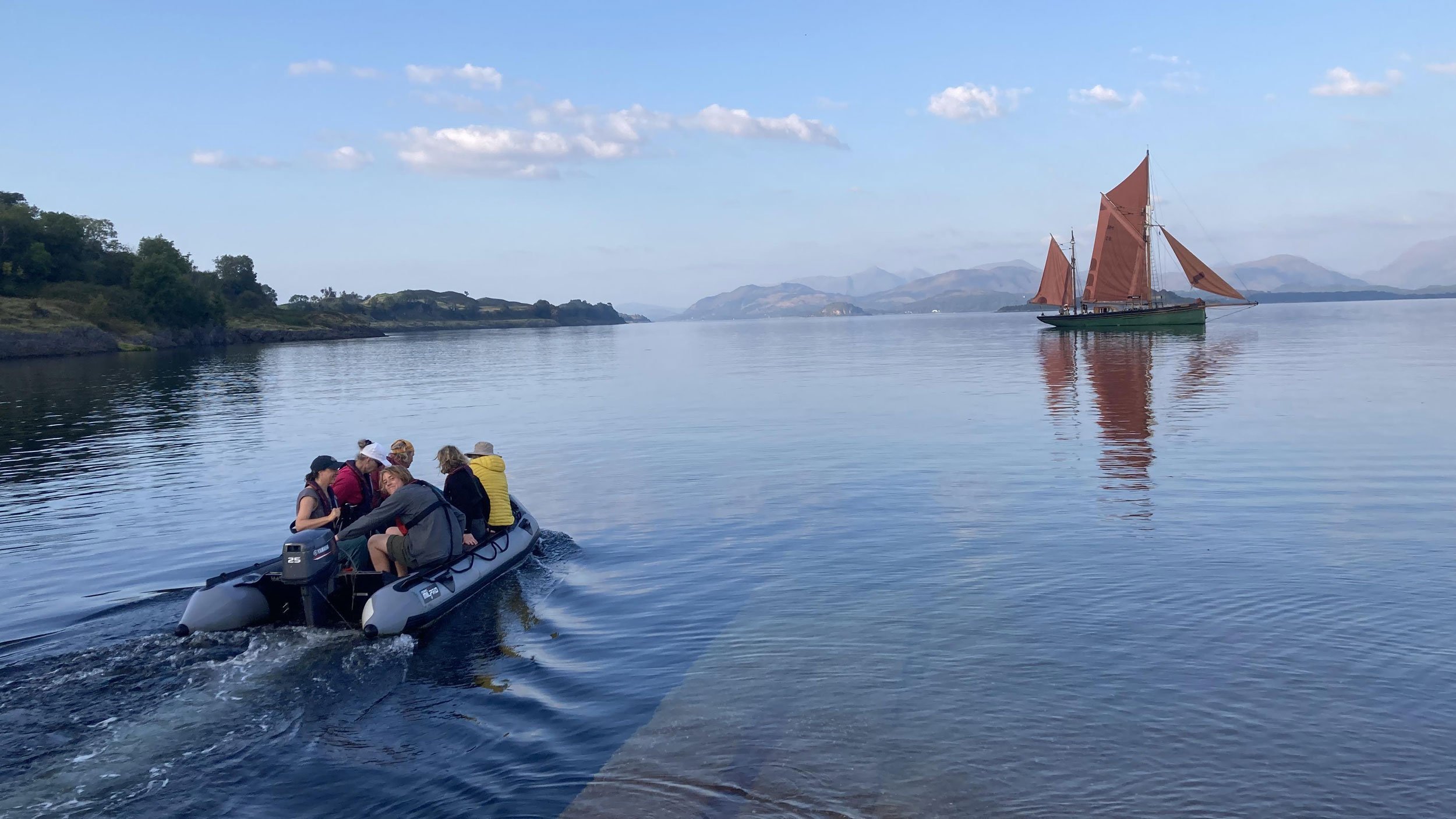 A group of people in a small inflatable boat on calm water near a shoreline, with a large sailboat with orange sails and mountains in the background.