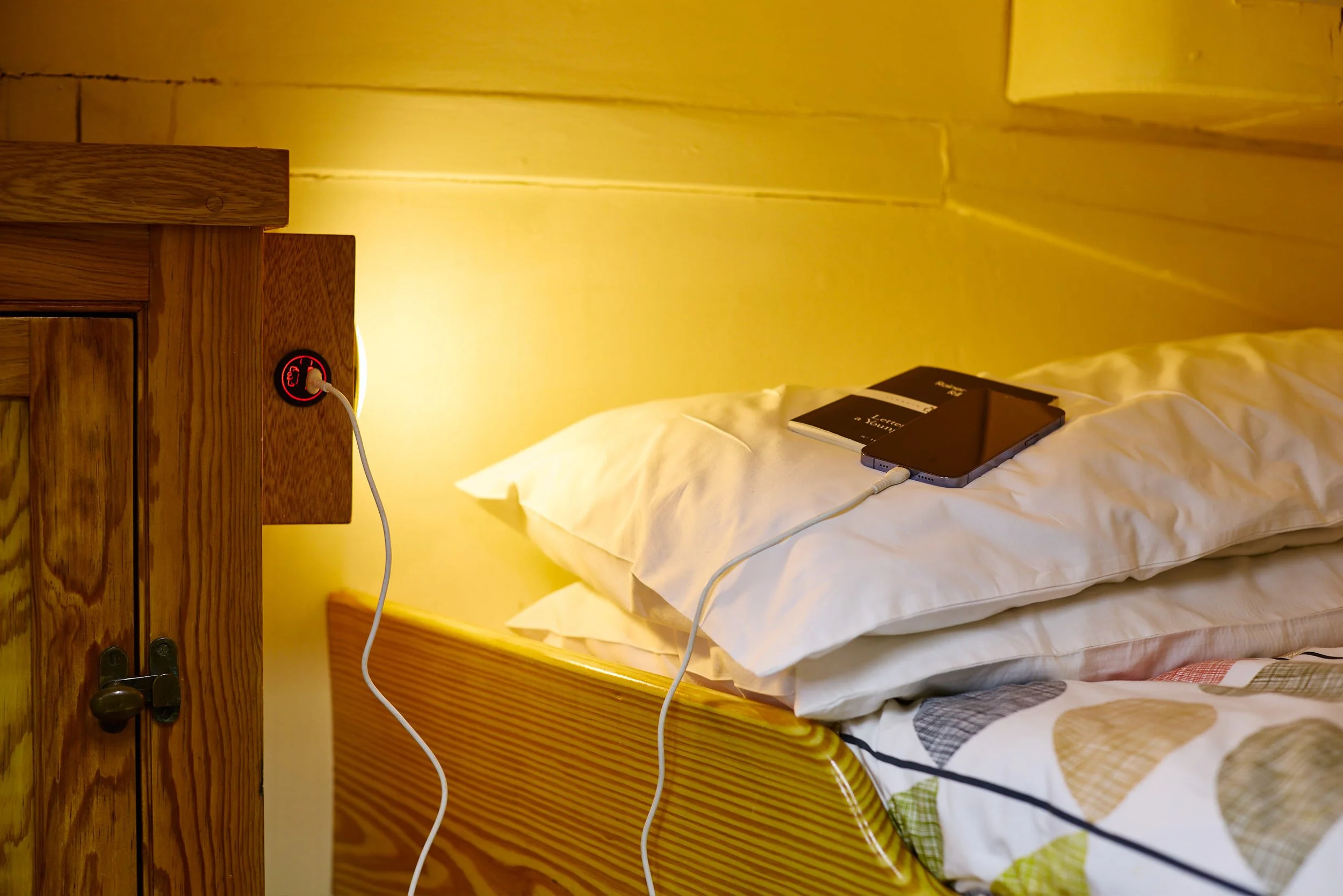 A bedroom with a bed and a pillow, a smartphone, a book, and charging cables plugged into a wall socket on the wooden bedside table. The room has yellow lighting and wooden furniture.