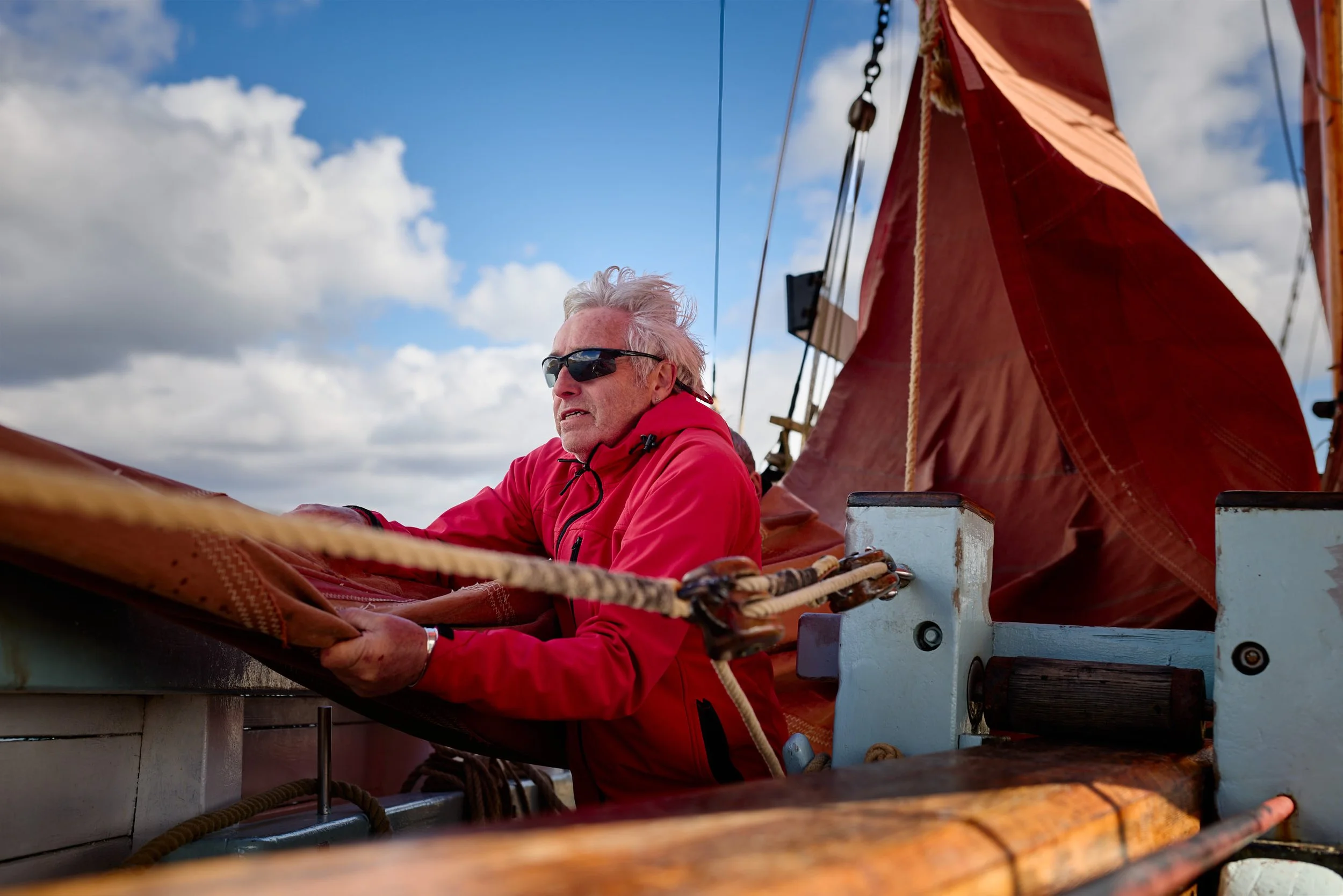 An older man with gray hair wearing sunglasses and a red jacket sitting on a boat with a red sail, looking out at the sky filled with clouds.