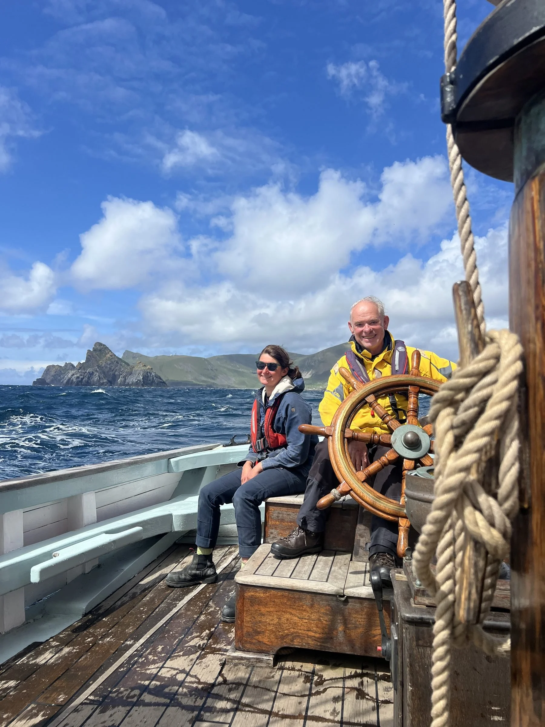 Two people sitting on Provident with a scenic coastal background, one smiling and the other wearing sunglasses, steering the boat with a ship's wheel, under a partly cloudy sky.