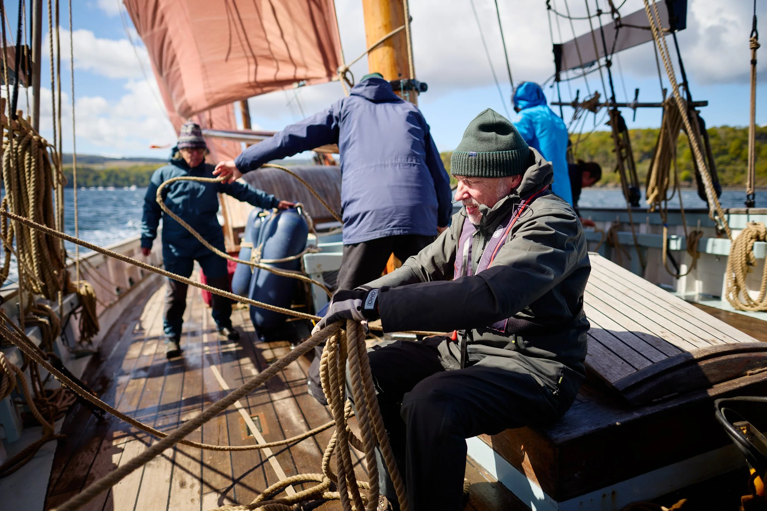 Older man wearing a gray jacket and green beanie working on ropes on the deck of a wooden sailing ship, with crew members in jacket and hood working nearby, water and trees in the background.