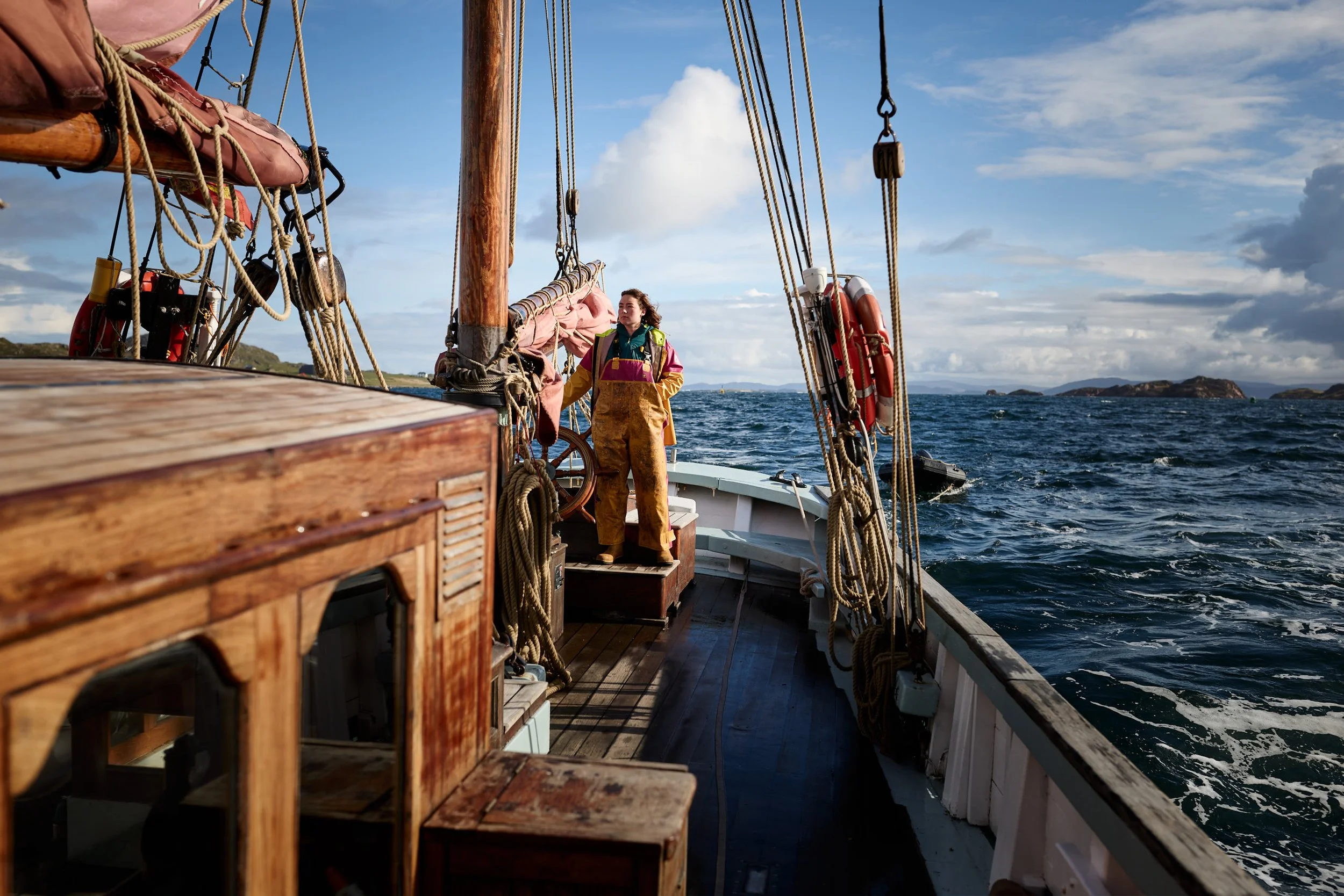 A person standing on the deck of a wooden sailing ship with tall masts, ropes, and rigging, looking out at the open ocean with a shoreline and hills in the distance under a partly cloudy sky.