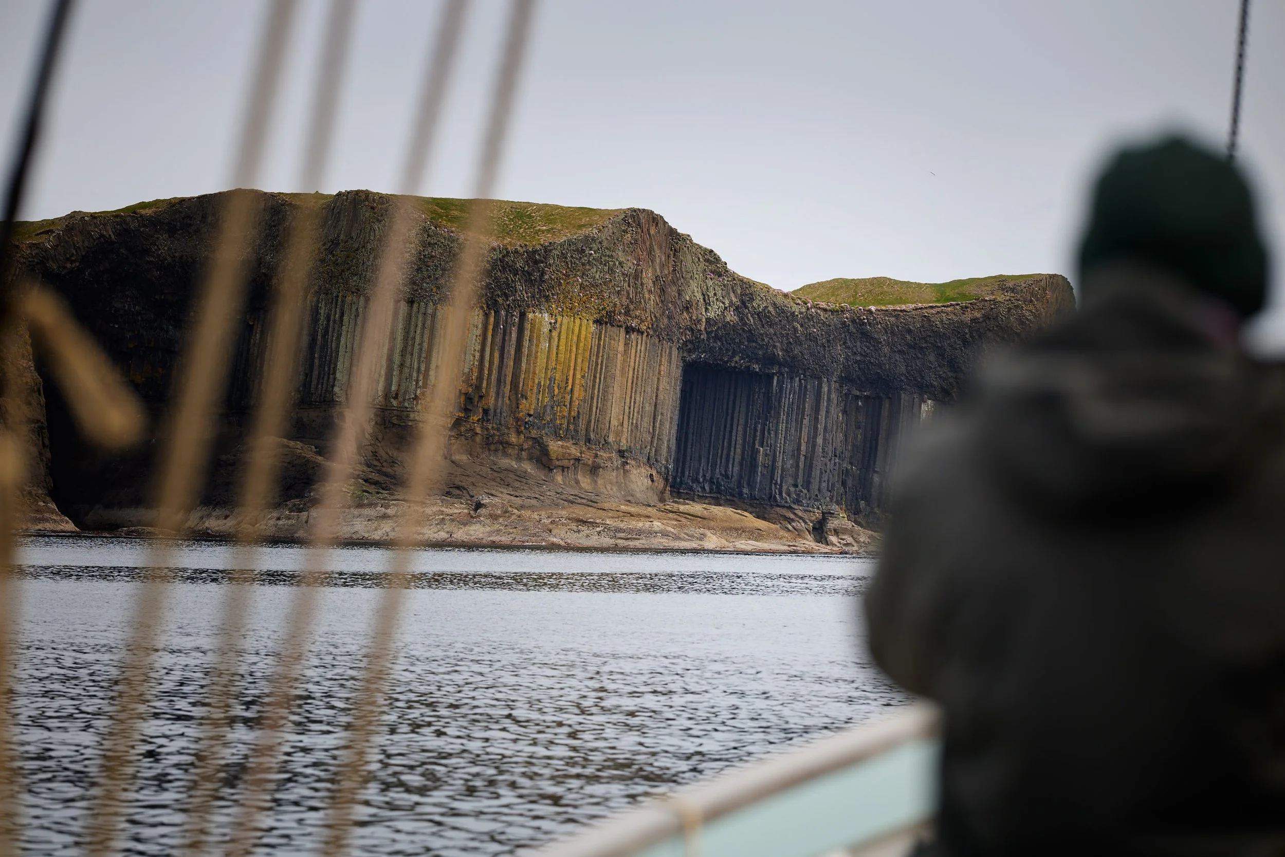 View of coastal cliffs with green patches, seen through a blurry mesh or cage, with water in the foreground and a gray, overcast sky.