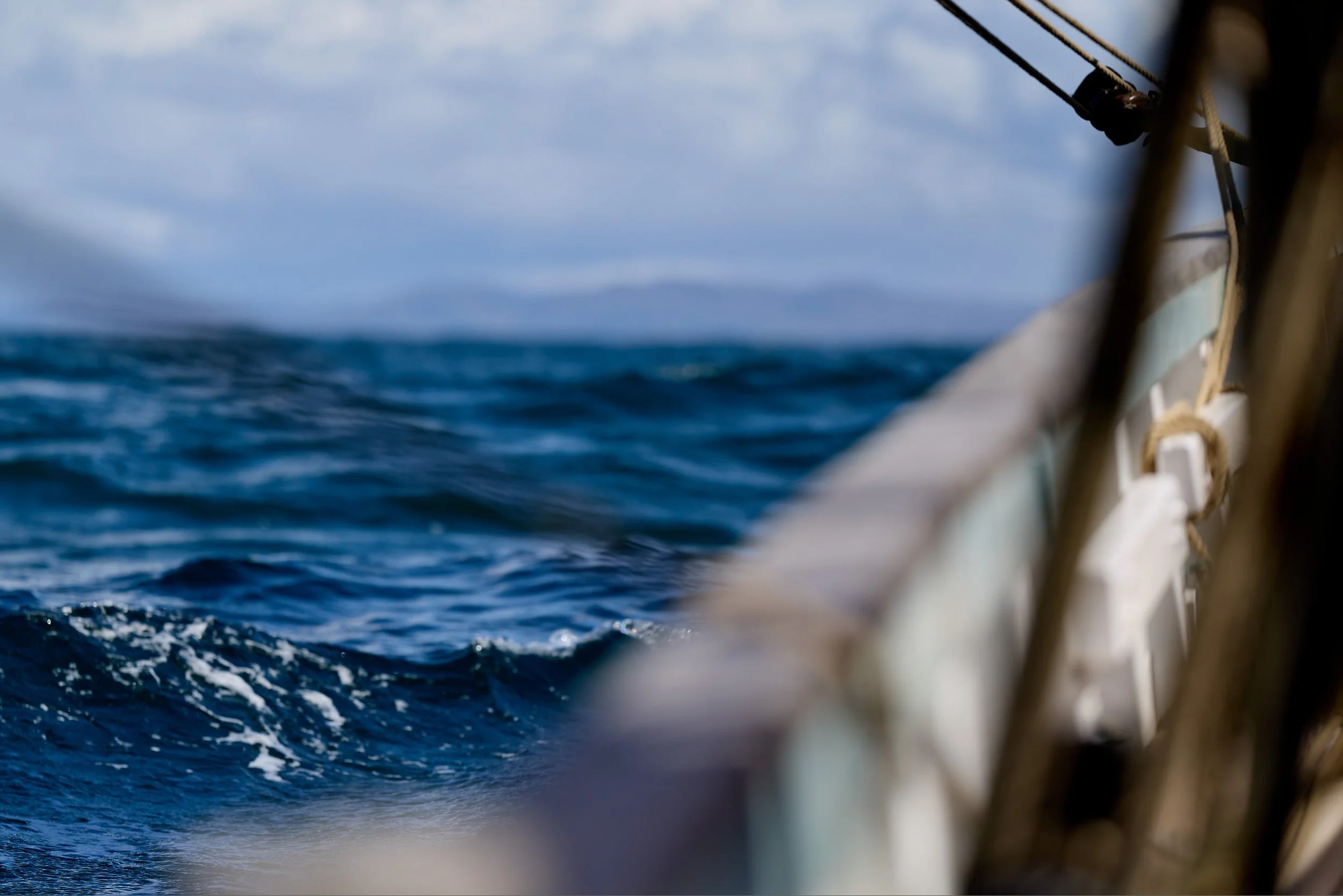 Close-up of ropes and rigging on a sailboat, with the ocean and cloudy sky in the background.