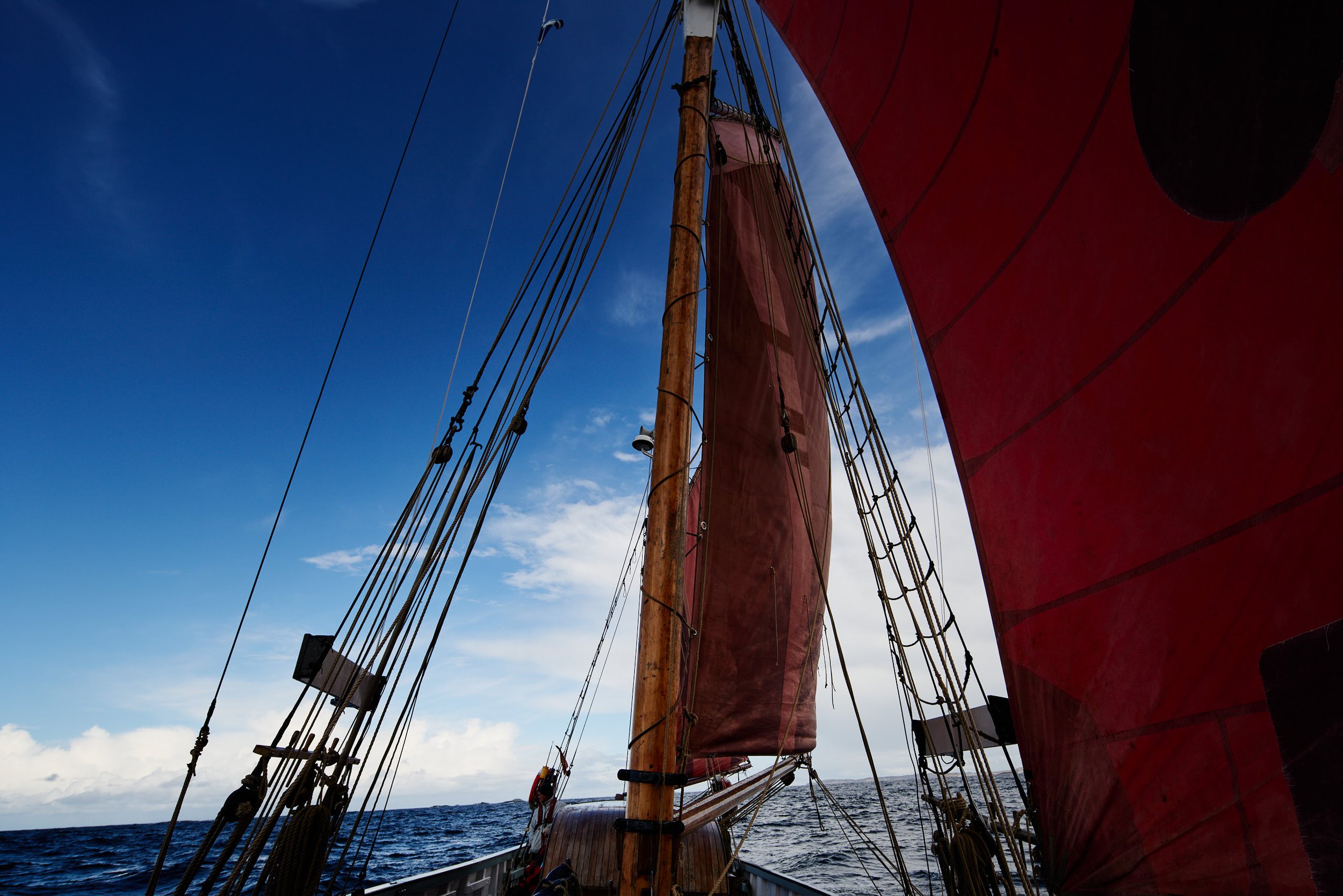 View from a sailing ship showing its mast, rigging, and red sails against a blue sky with some clouds, on open ocean water.