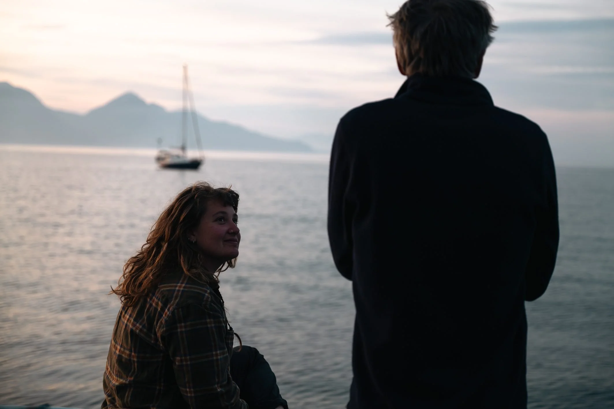 A woman with long brown hair and a man with short dark hair stand near the water, with a boat and mountains in the background during sunset.