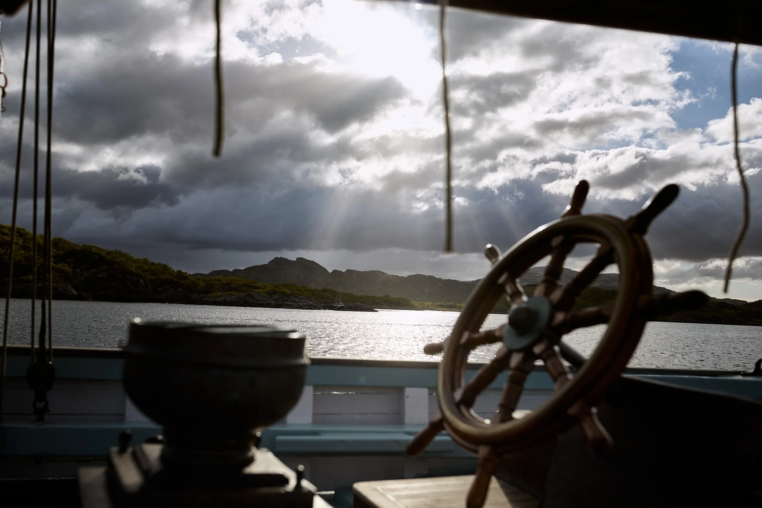 View from a boat or dock showing a water body with a sailboat, mountains in the background, dark clouds in the sky, and a steering wheel and fishing gear in the foreground.