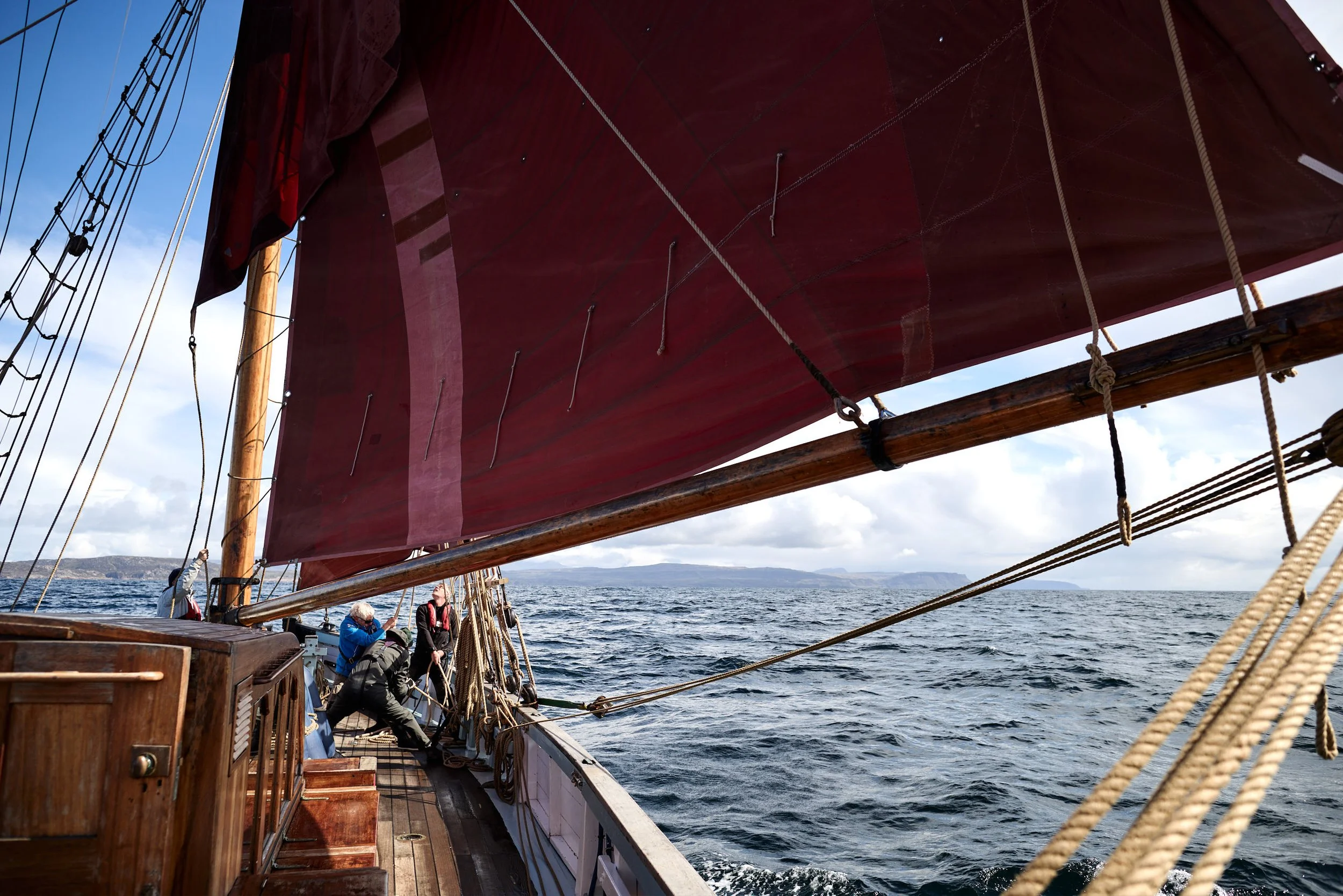 People on a wooden sailboat with large red sail, ropes, and rigging, sailing on water under a partly cloudy sky with land in the distance.