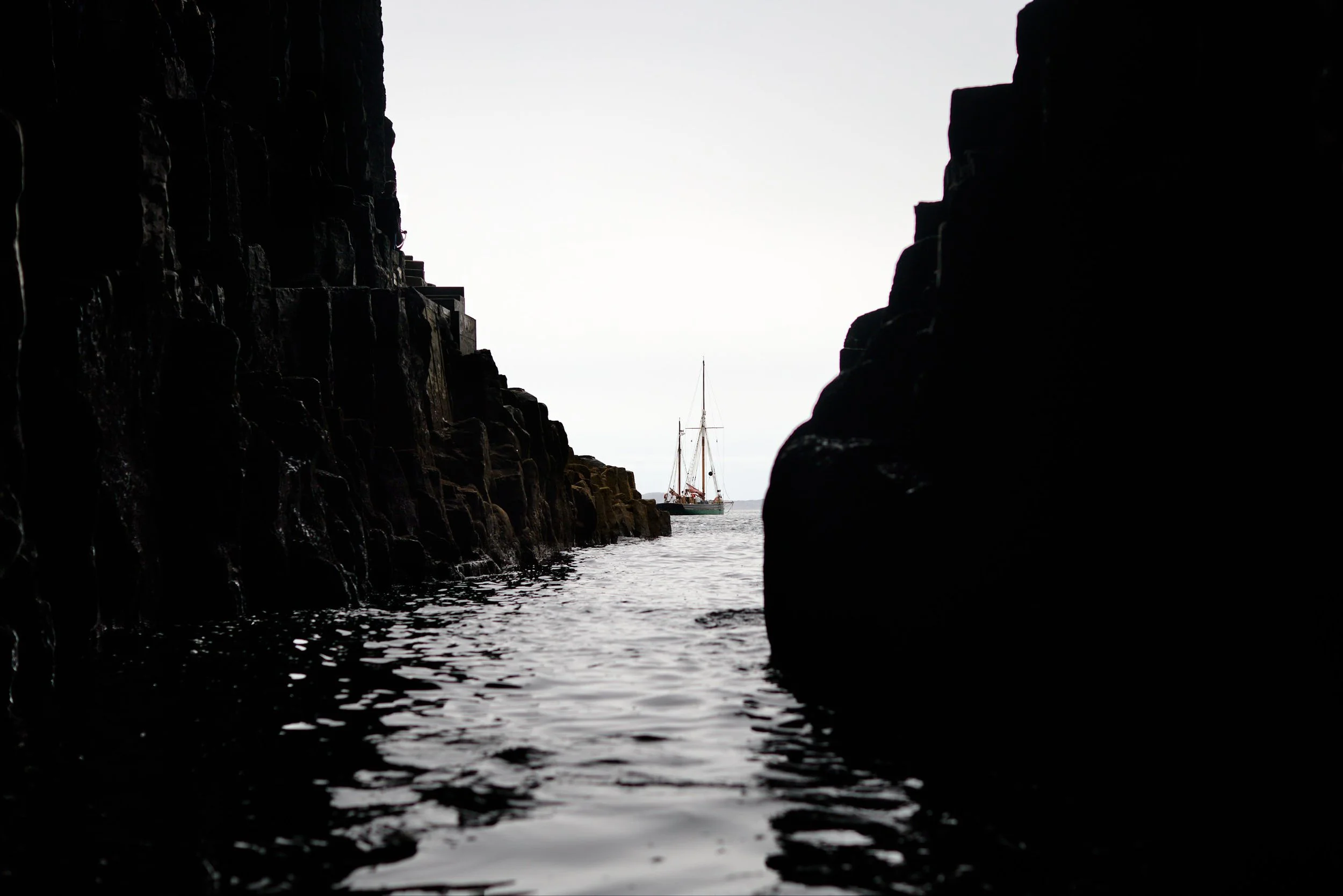 A sailboat is seen through a rocky coastal crevice with dark rocks framing the scene, and the sky is overcast.