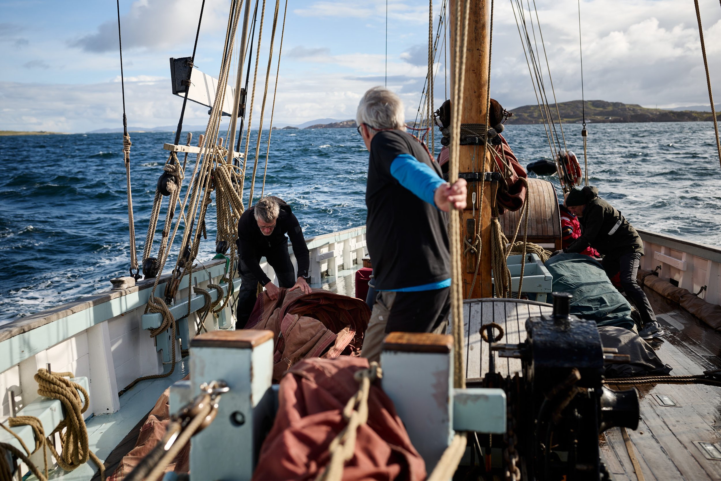 People on a boat with ropes, wooden masts, and a view of water and distant islands or land.