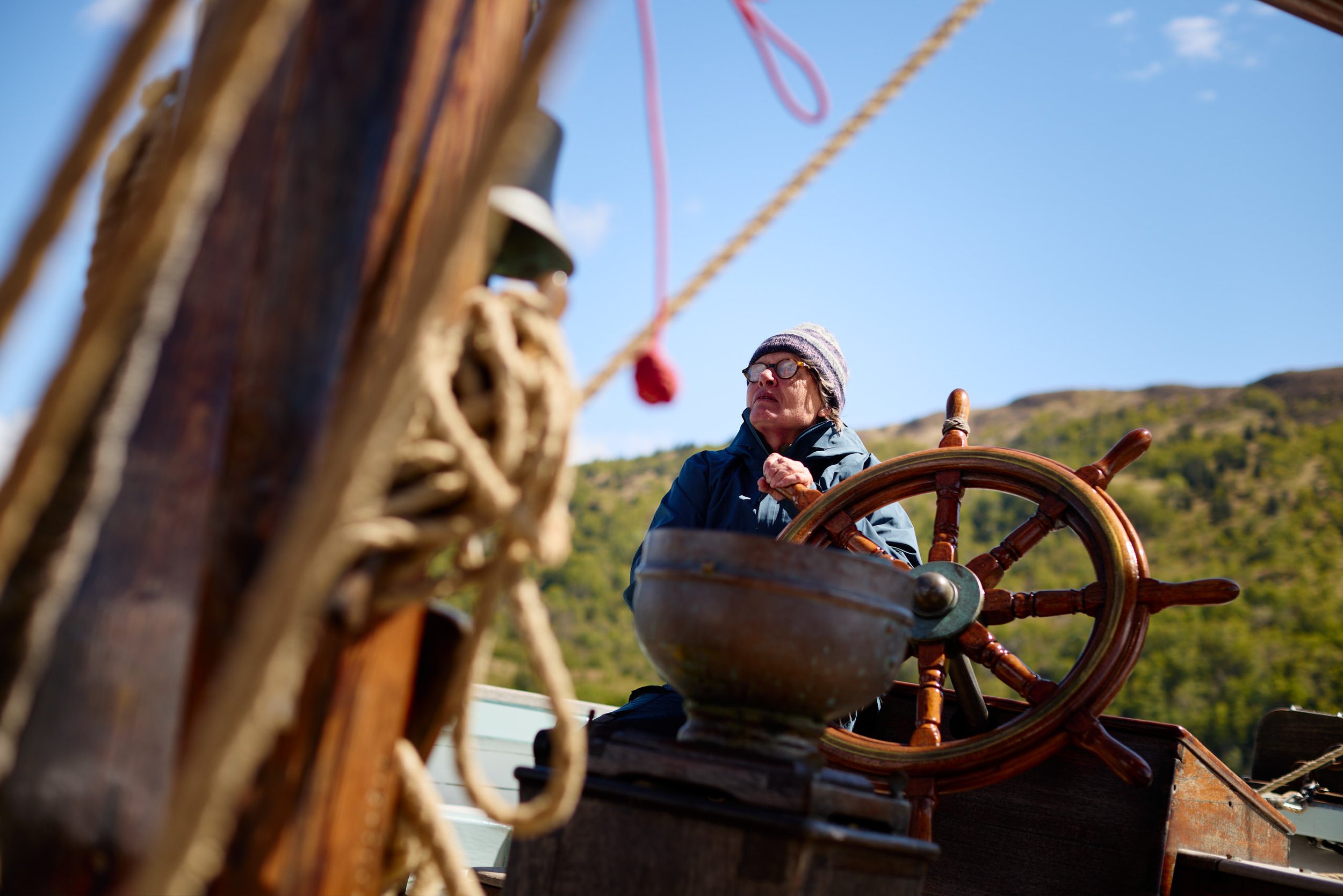 A person wearing glasses, a knit cap, and a jacket is steering a wooden ship at sea, with scenic hills and a blue sky in the background.