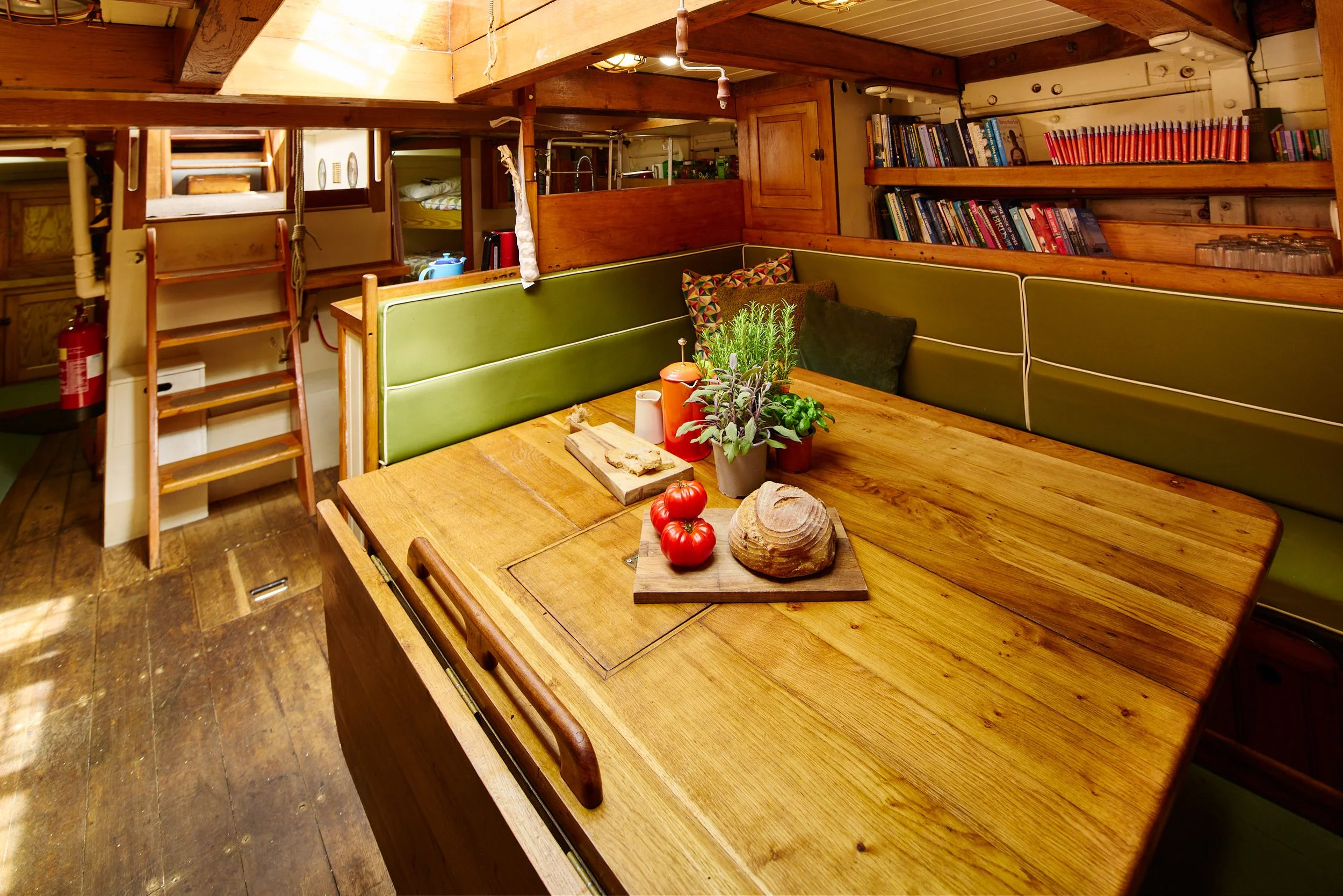 Wooden table with a potted plant, tomatoes, and other small items, with green cushioned seating around it in a cozy wood-paneled room filled with books and kitchen items.