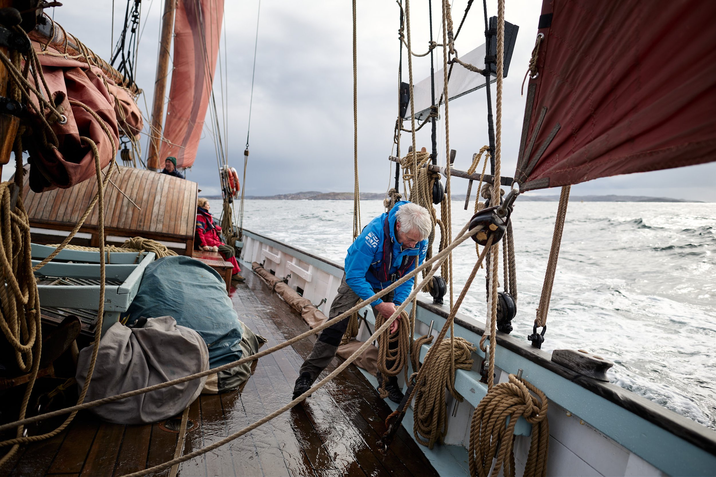 People working on a sailing ship's deck in rough weather, surrounded by ropes, sails, and equipment with water and distant land in the background.