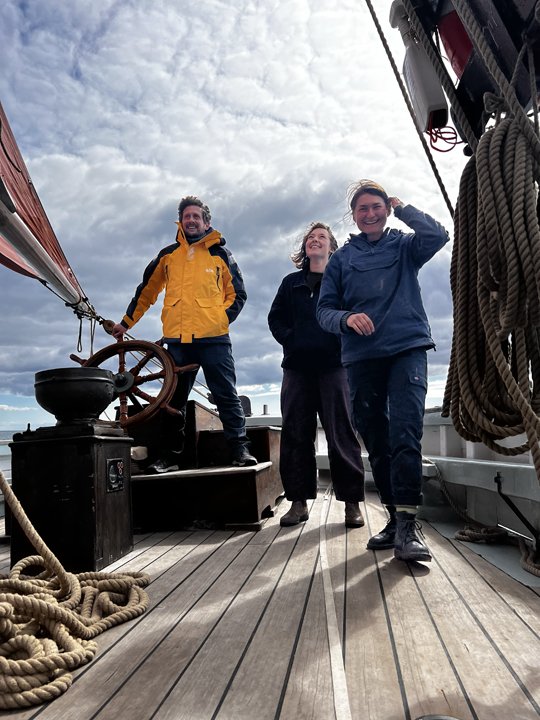 Three people standing on the deck of a sailboat, smiling and enjoying the weather, with cloudy sky above.