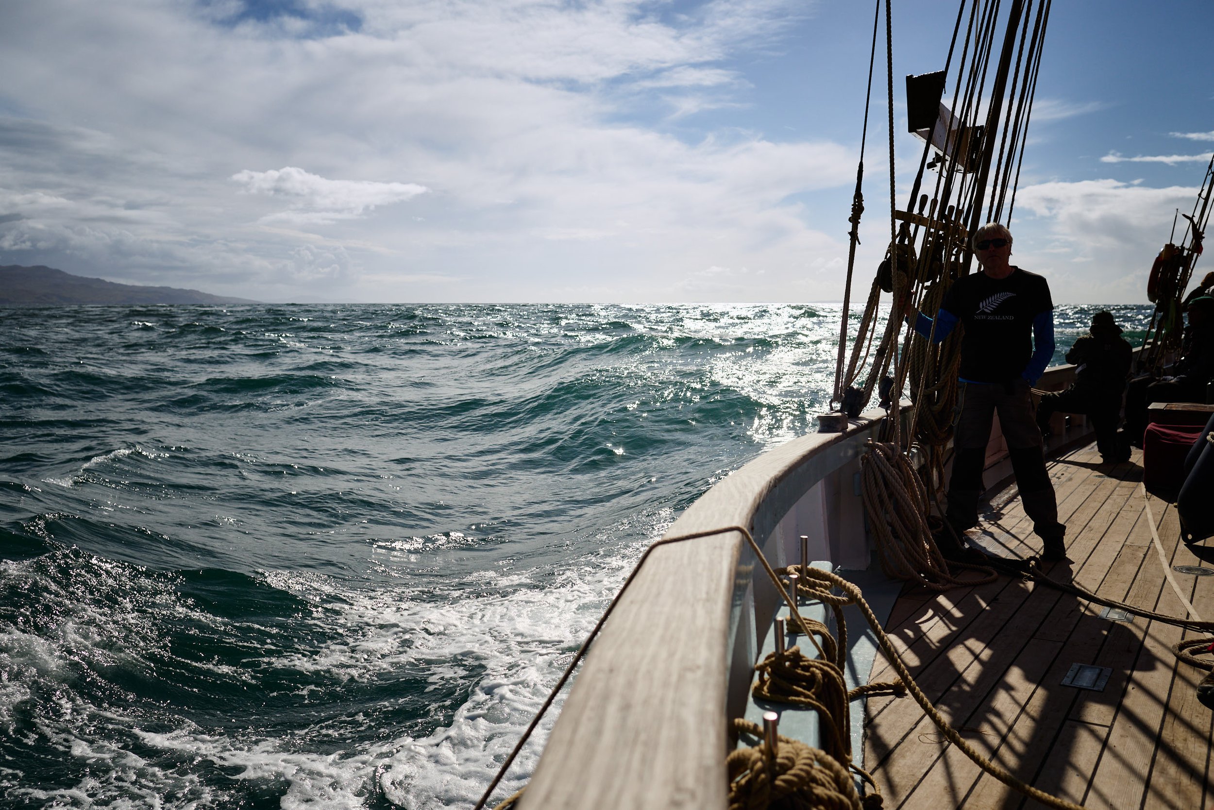 A person standing on the deck of a boat with sailing rigging, overlooking the ocean with waves and a distant landmass under a cloudy sky.