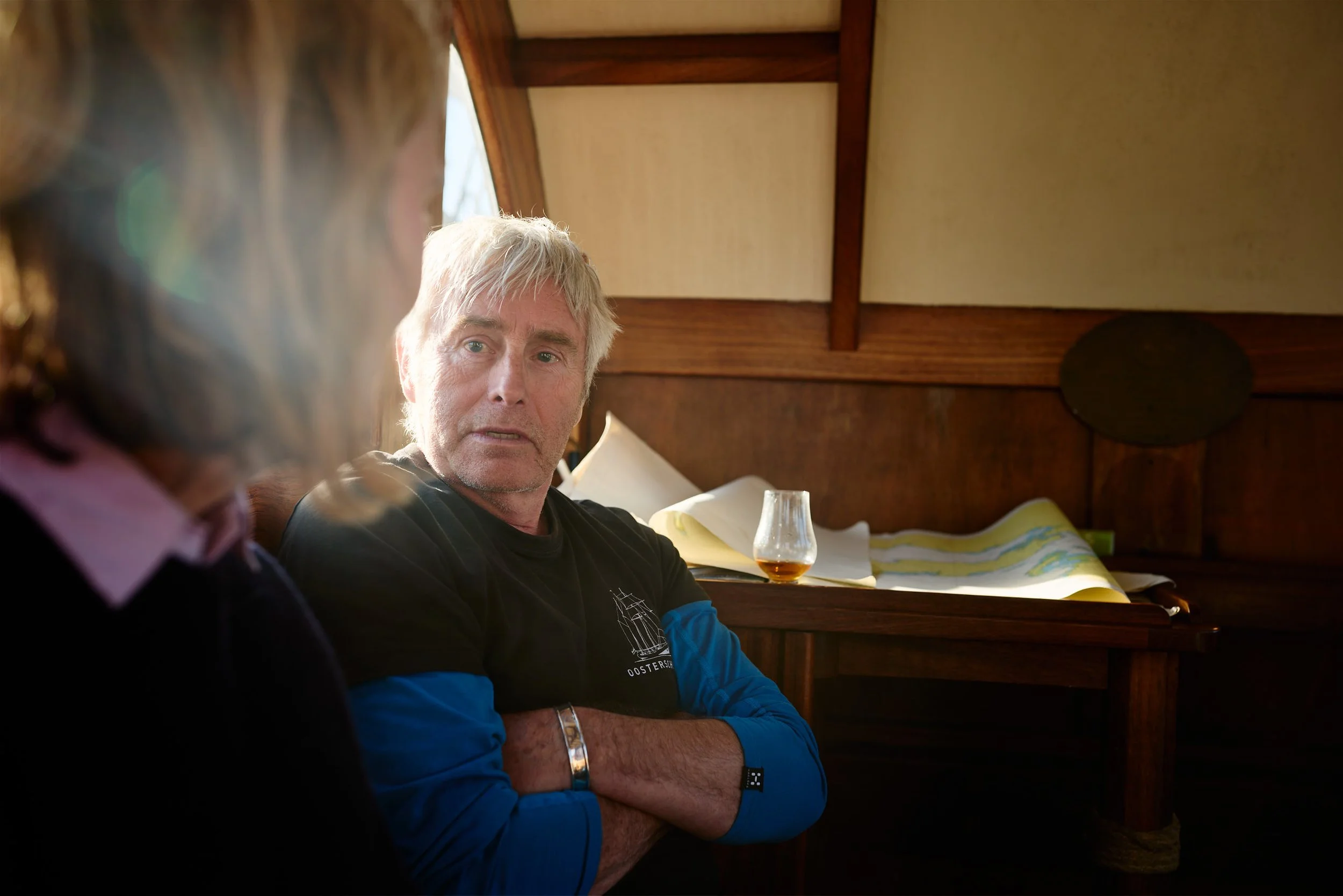 A man with gray hair and a beard looks at a woman with brown hair in a cozy room with wooden decor. There is a glass of dark beverage and a map or book on the bed in front of him.