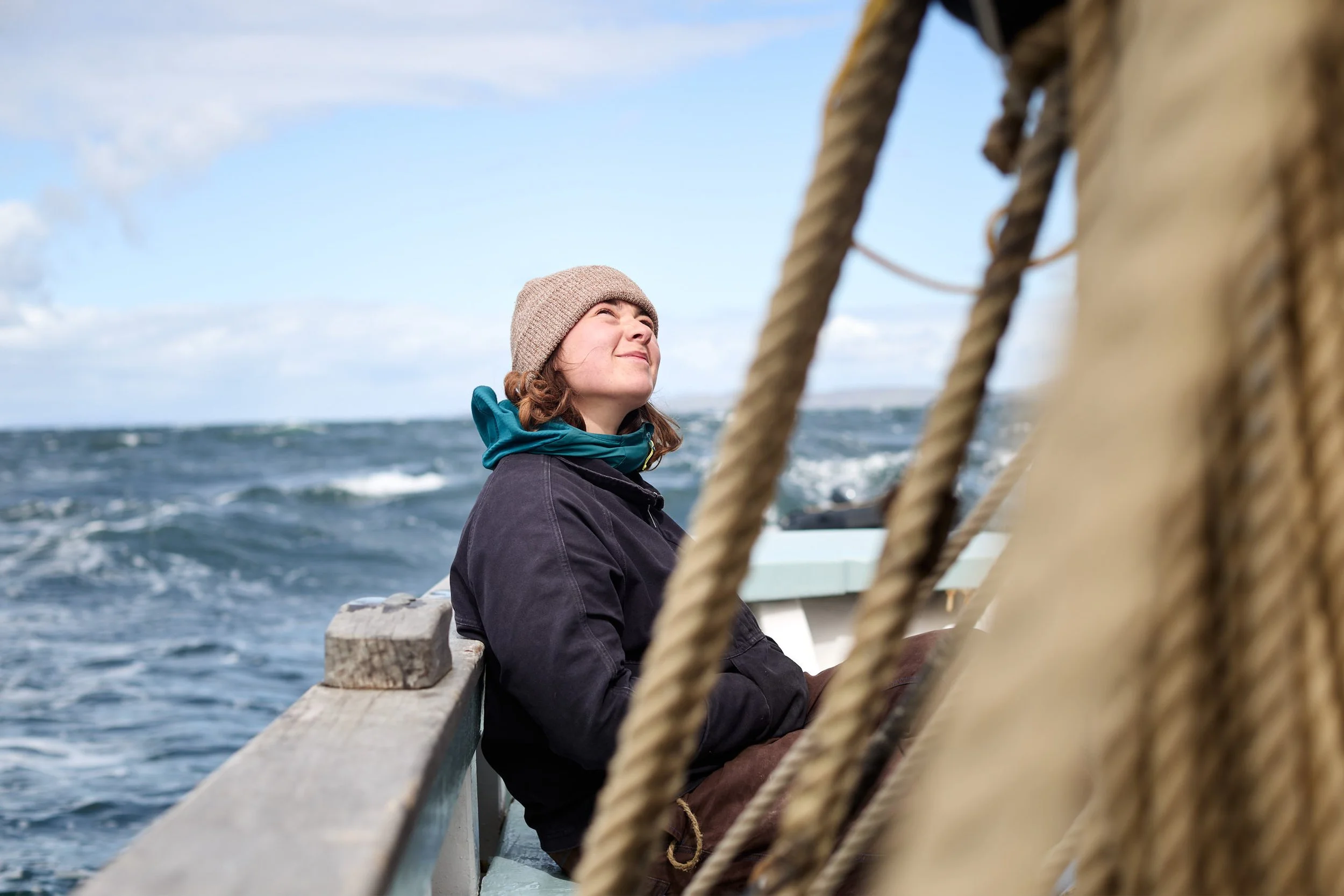 A woman wearing a pink knit hat and a dark jacket is sitting on a wooden bench on a boat, with the ocean and blue sky in the background. She appears to be enjoying the view or the moment.