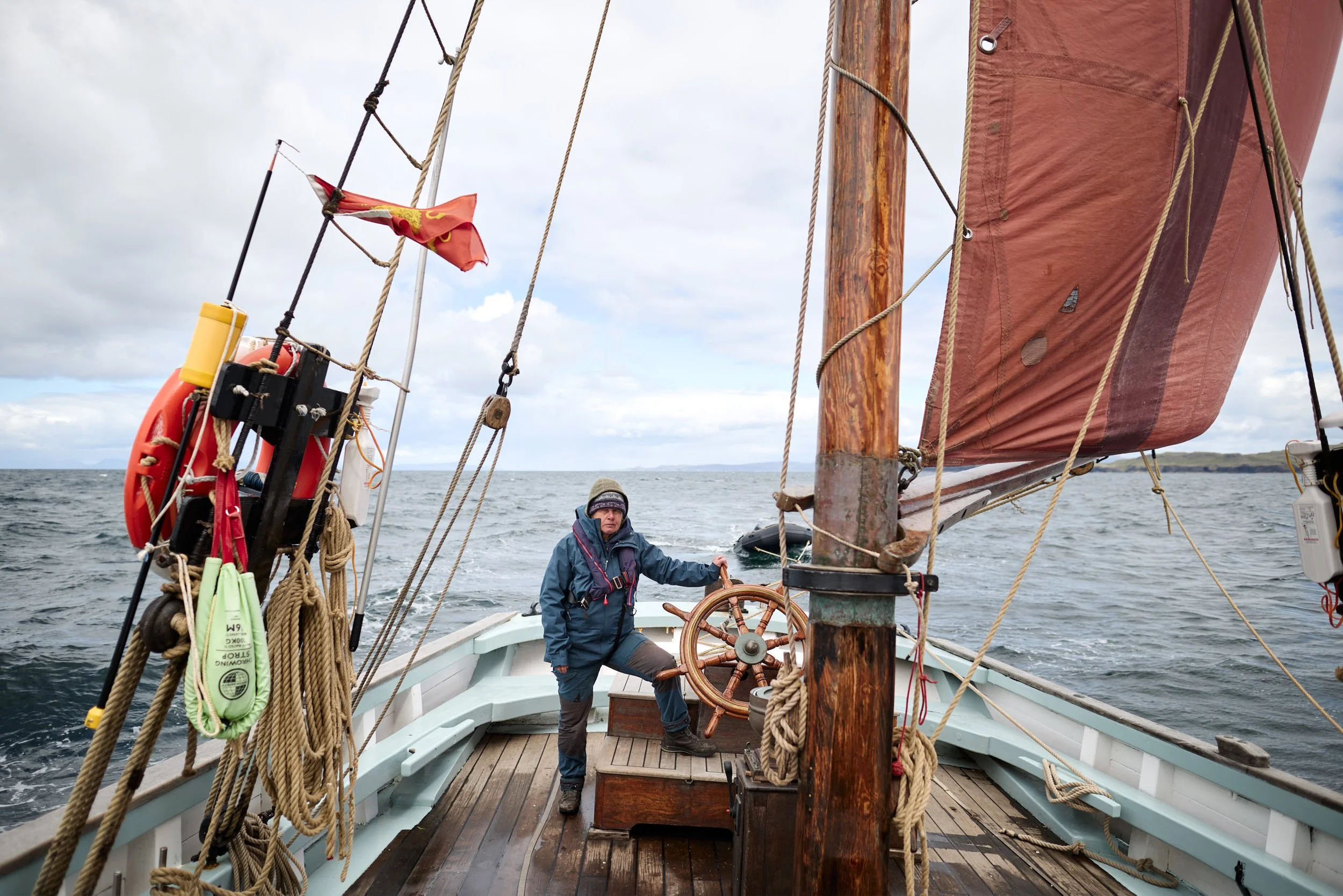 A person steering a sailboat on the water, with the boat's wooden mast, ropes, and red sail visible. The person is dressed in a blue jacket and gray pants, wearing a hat, and standing at the helm.