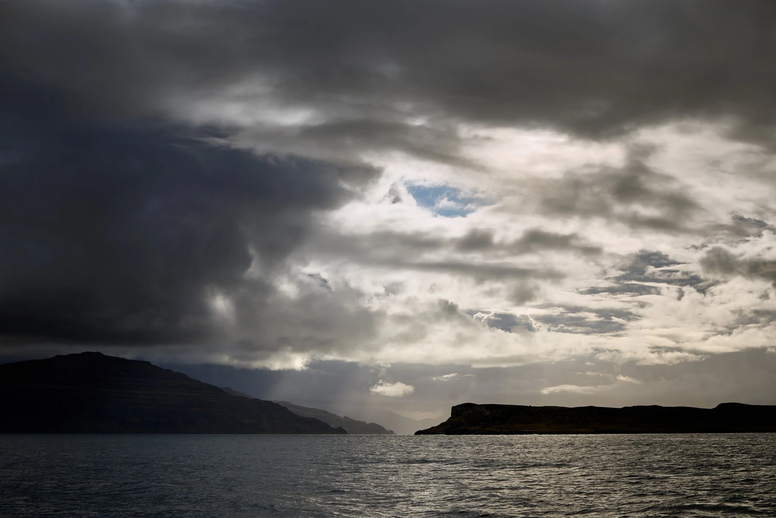 Cloudy sky over a large body of water with silhouettes of hills or mountains in the distance.