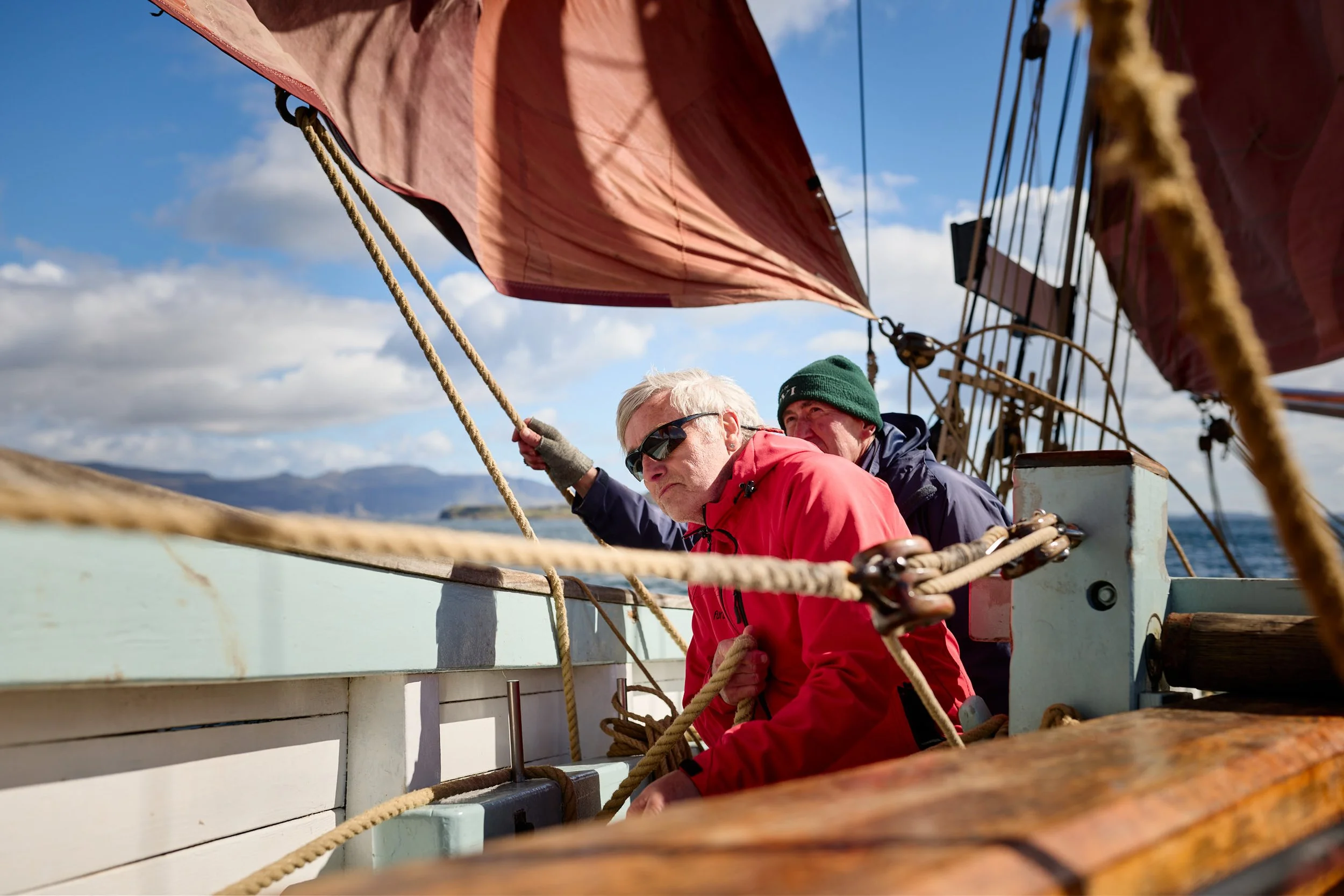 Two men sailing on a boat in a bright, partly cloudy day with distant mountains in the background. One man with white hair and sunglasses is handling a rope, while the other man wearing a green beanie looks on. The boat has red sails and wooden raili