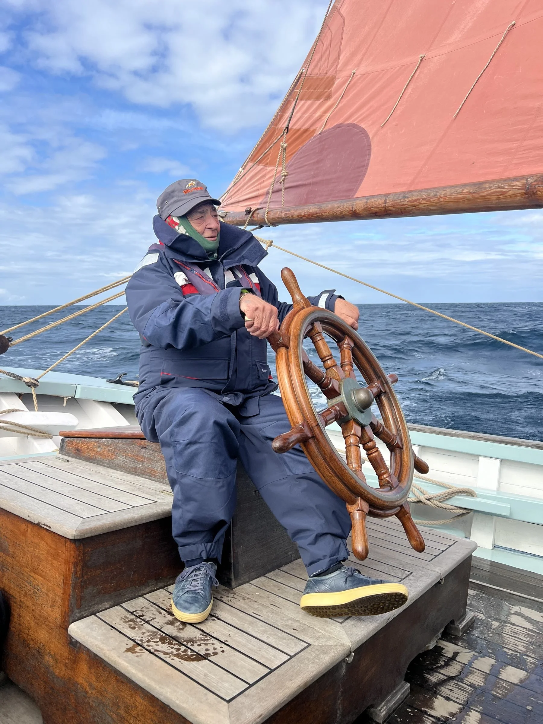An elderly man wearing a jacket and hat is steering a wooden ship's wheel on a boat sailing in the ocean, with sail and sky visible in the background.