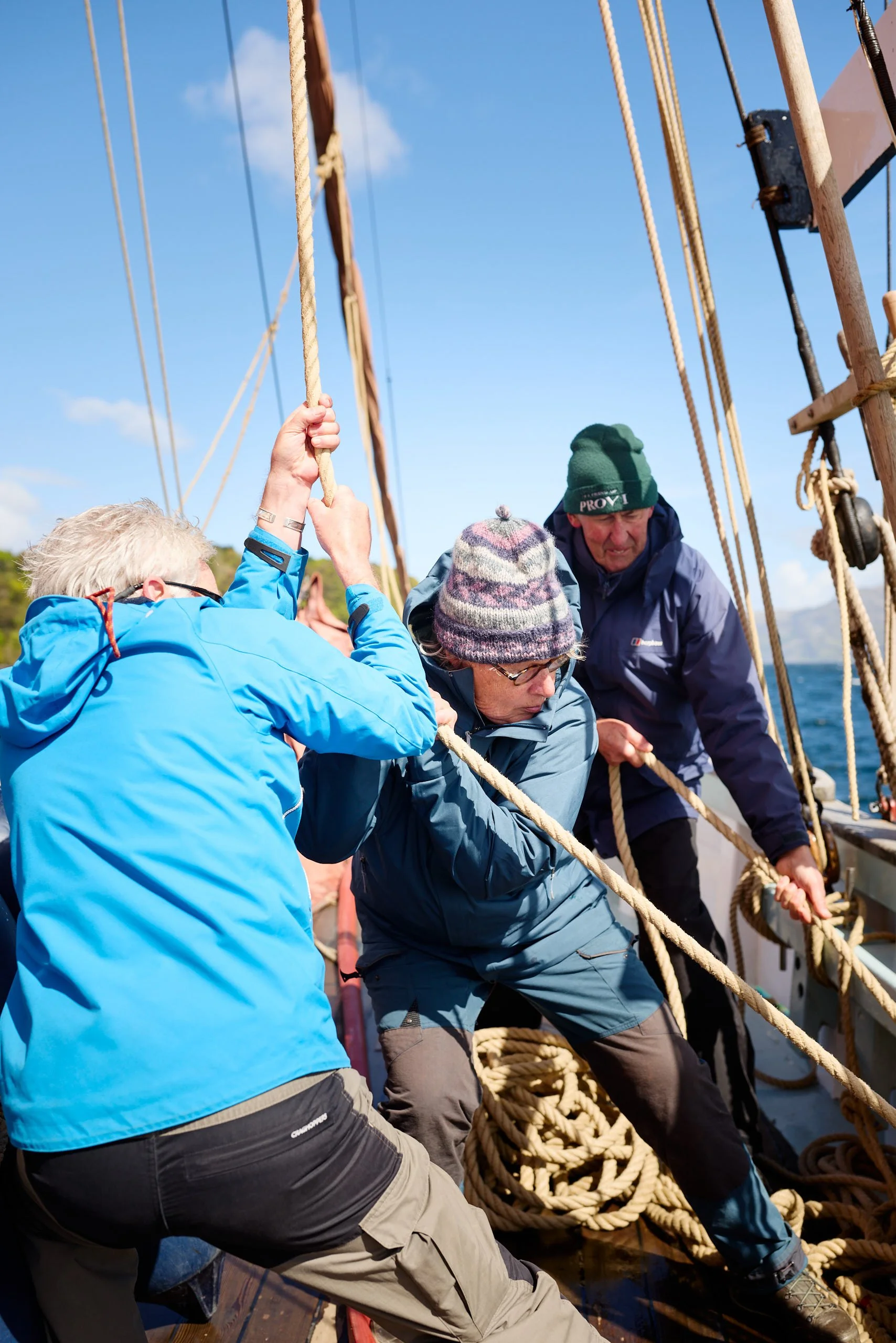 People on a sailboat pulling on ropes, with a clear blue sky in the background.