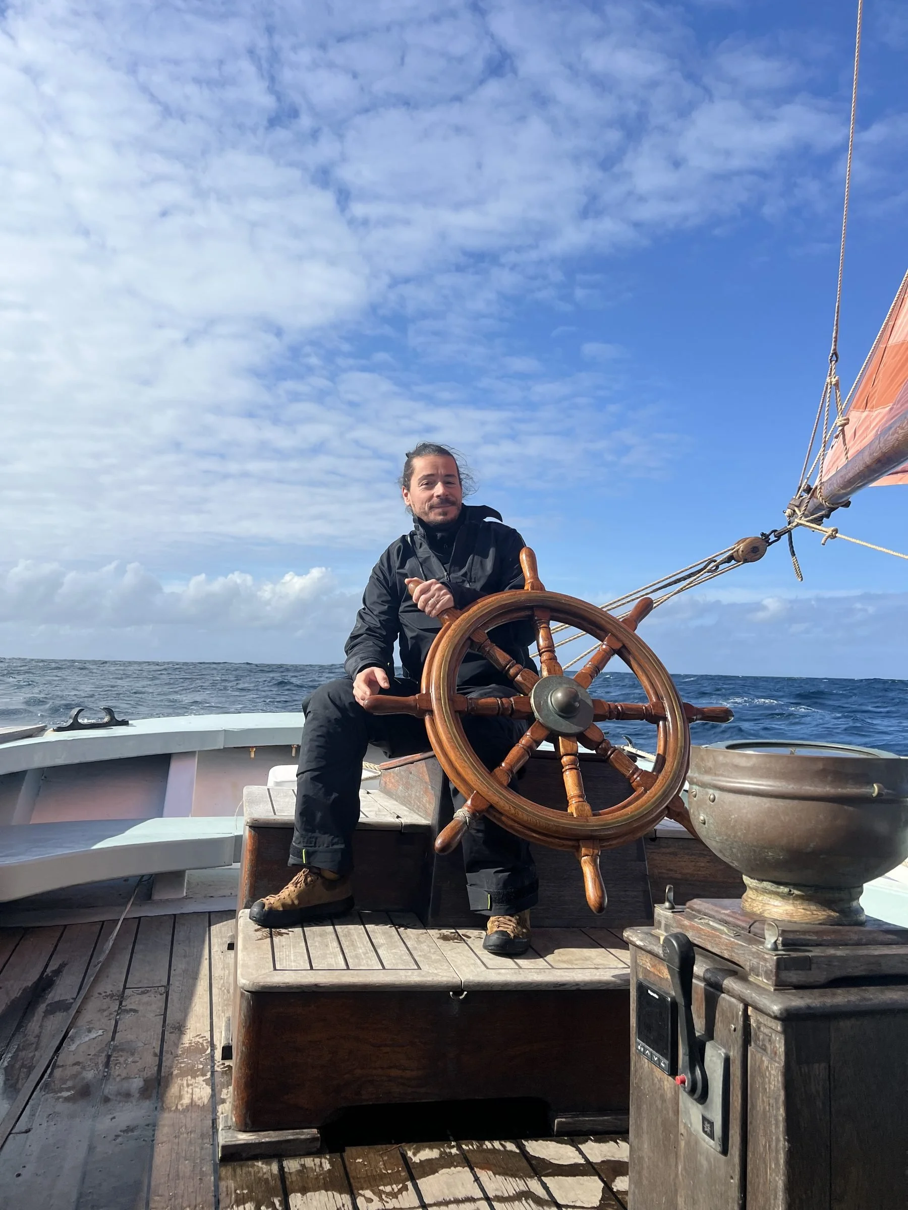 A man sitting at the steering wheel of Provident on the ocean, with a blue sky and scattered clouds above.