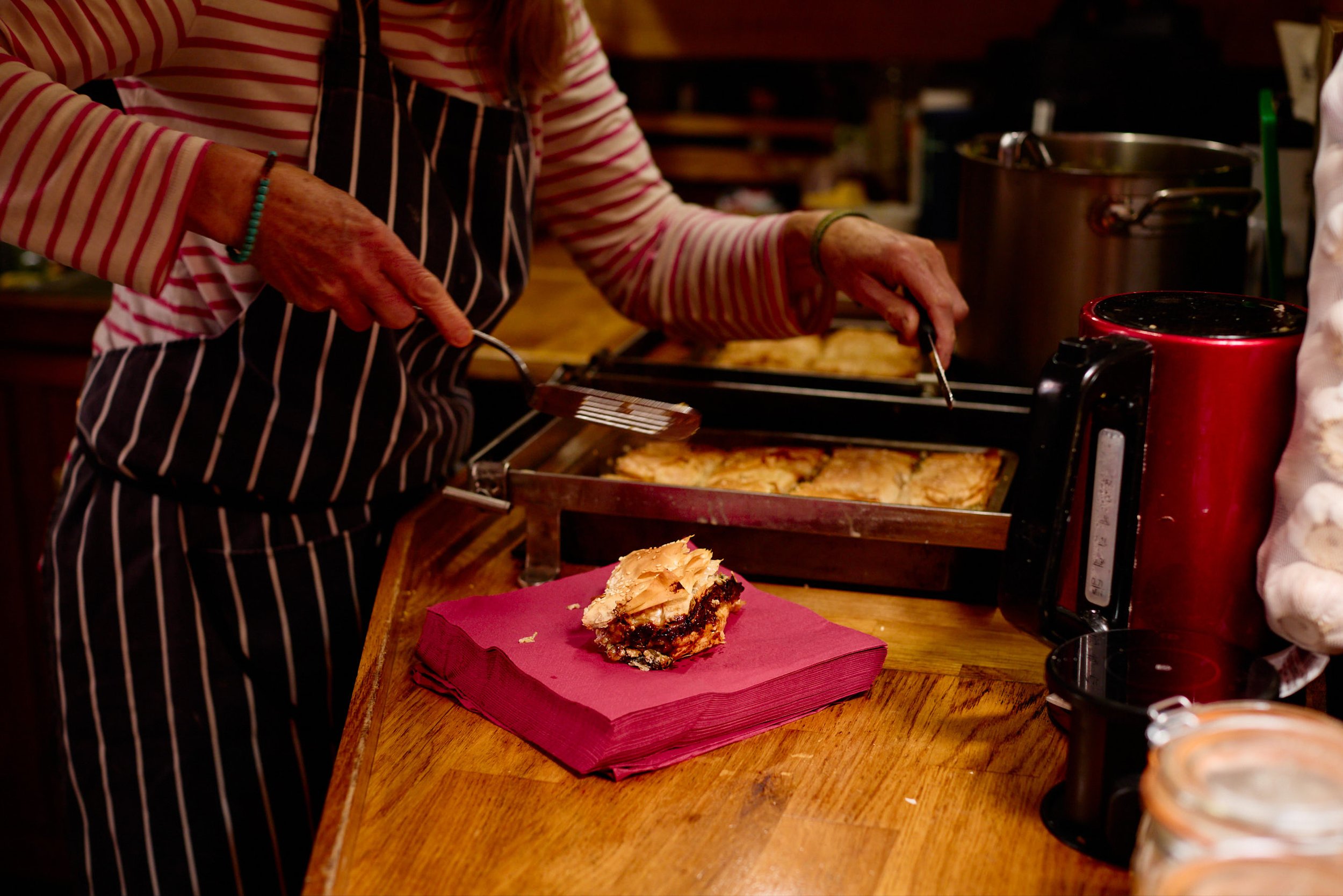 A person wearing a striped shirt and apron slicing a pie on a wooden counter, with a slice of pie on a red napkin in the foreground and various kitchen appliances and utensils around.