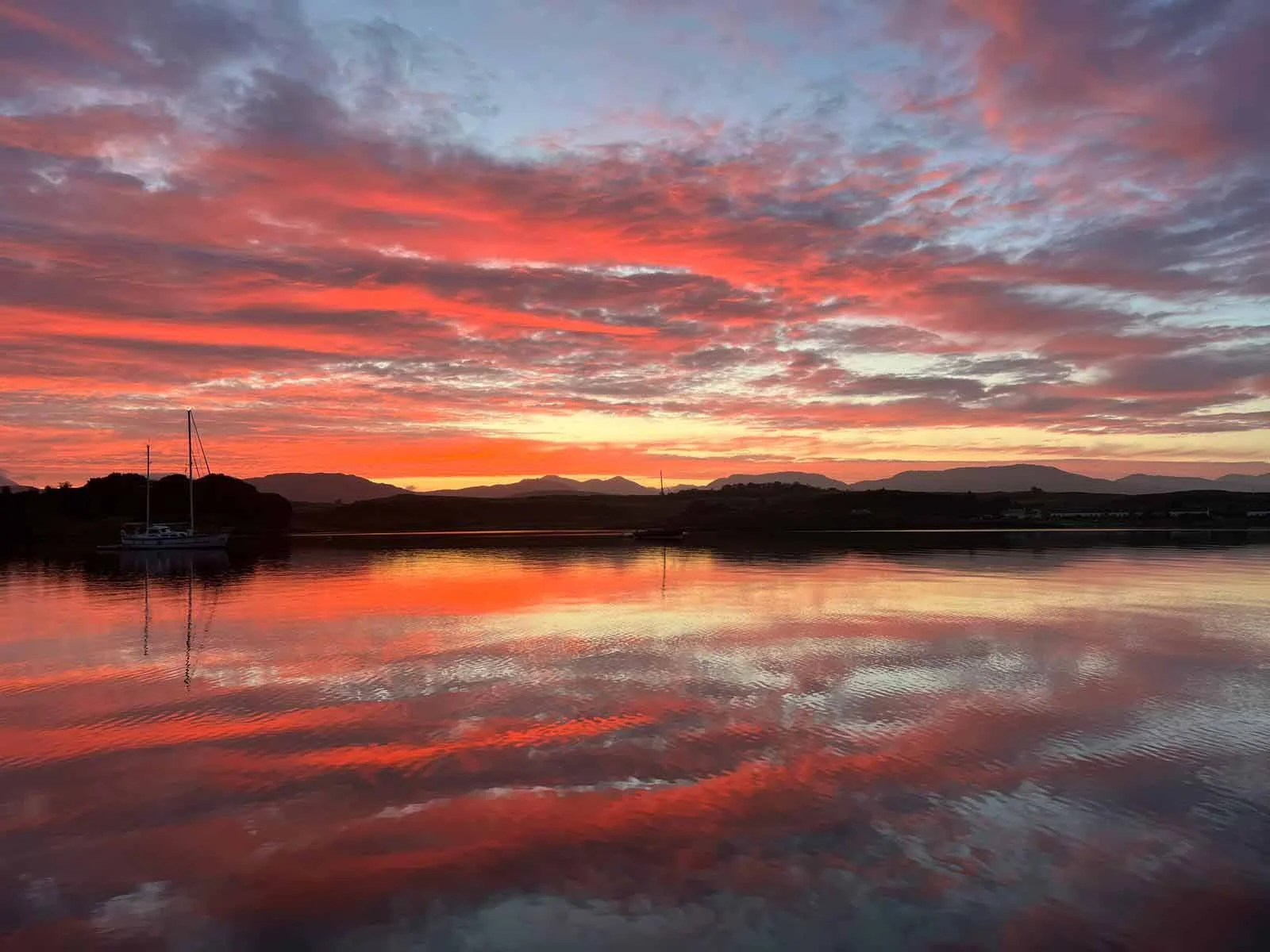 A scenic view of a sunset over a calm body of water with a sailboat on the left, silhouetted trees, distant hills, and a colorful sky with shades of pink, orange, and purple reflected in the water.