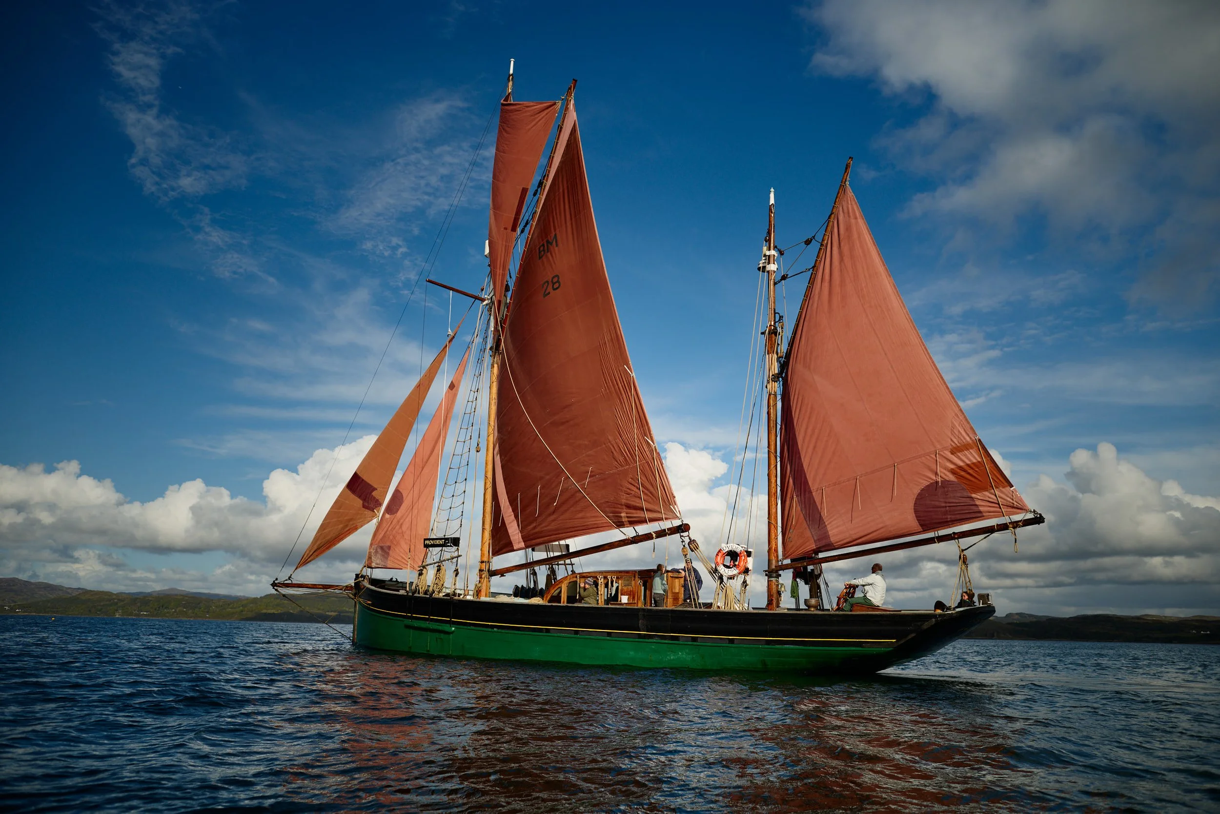 Tall ship, Provident, with reddish-brown sails and a green hull on the water, under a partly cloudy sky.