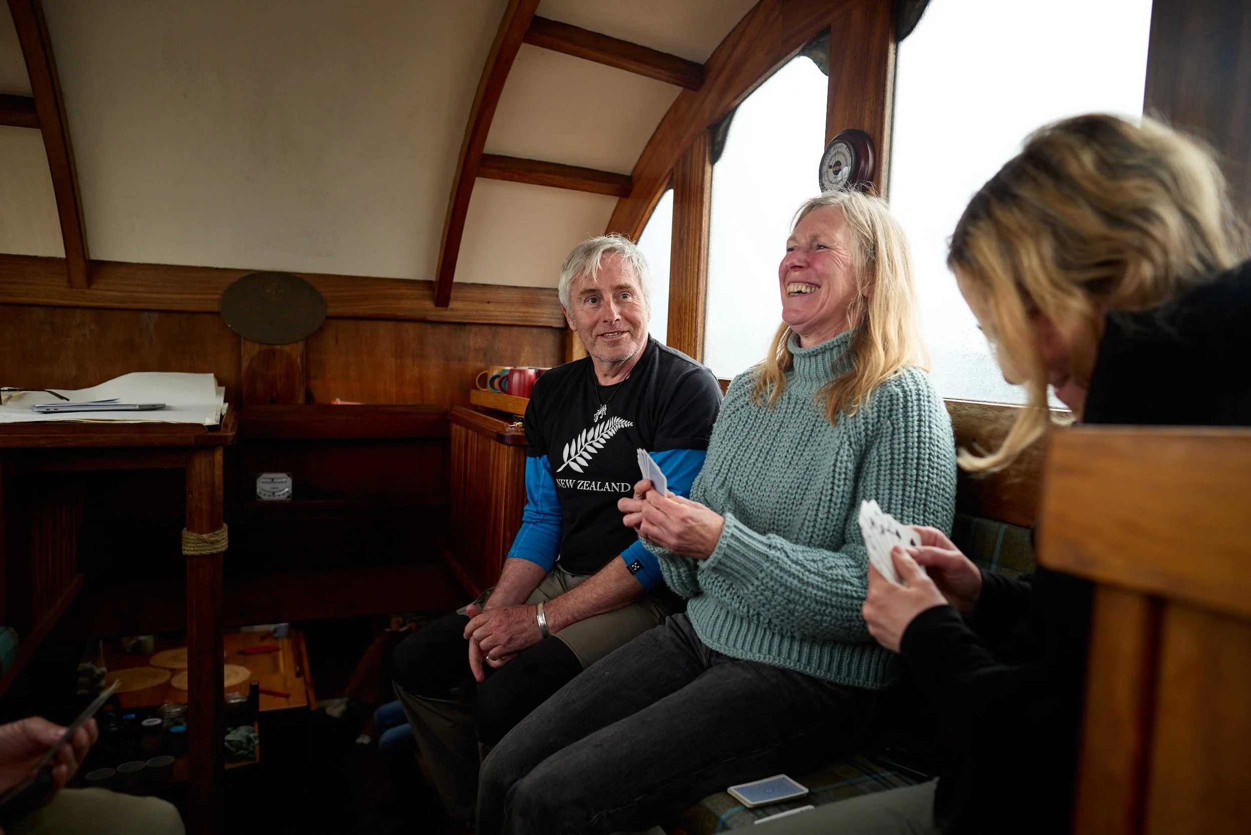Three adults sitting on a wooden bench inside a cozy room, smiling and engaging in conversation, with a person partially visible on the right holding playing cards.