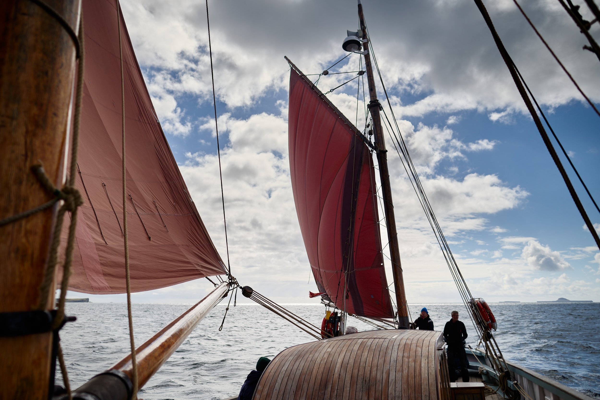 View from the deck of a sailboat looking up at the mast with red sails against a partly cloudy sky. People are visible on the boat, near the water.