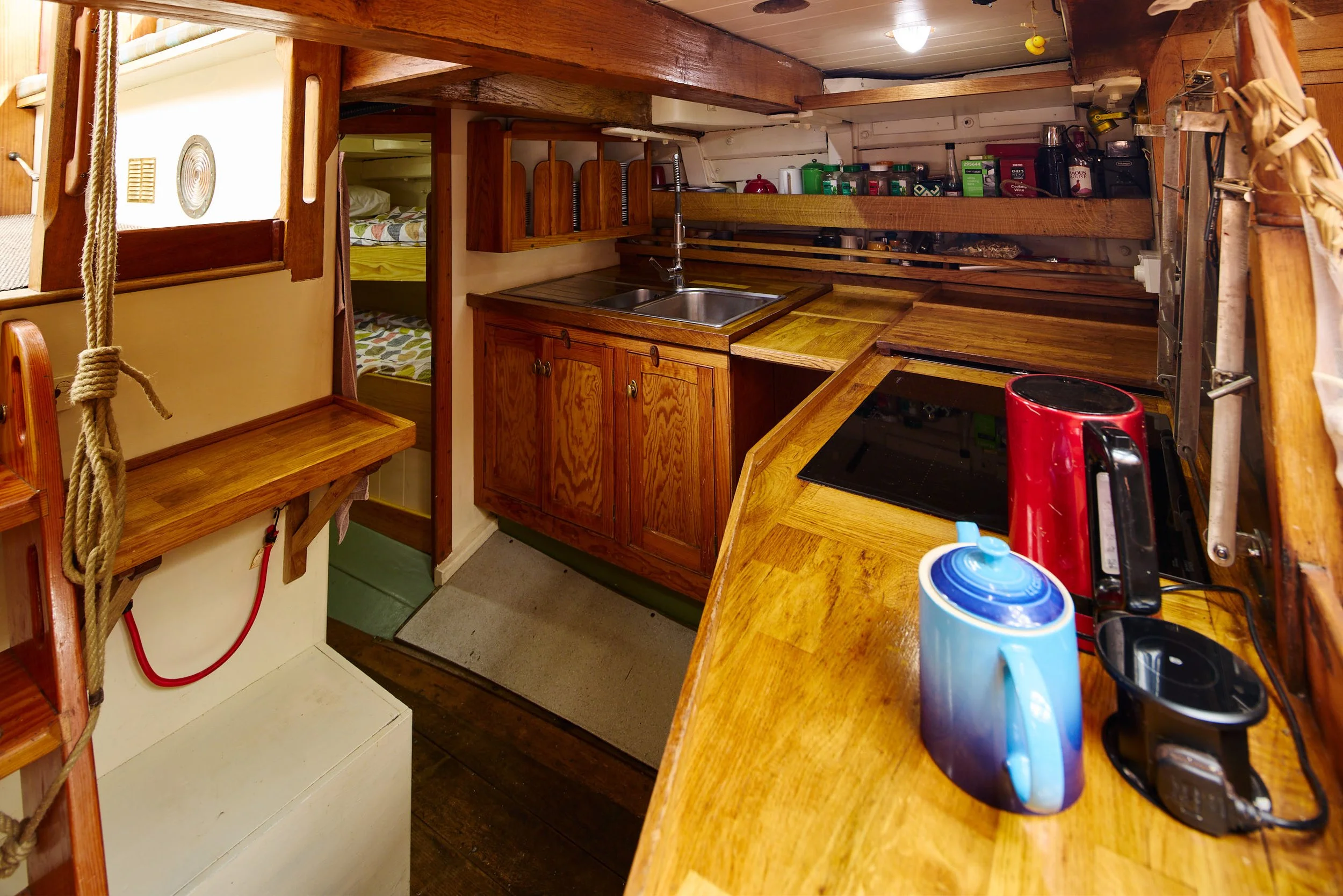 A compact kitchen with wooden cabinets, a sink, and various kitchen items on shelves, with stairs leading to a sleeping area with bunk beds visible in the background.