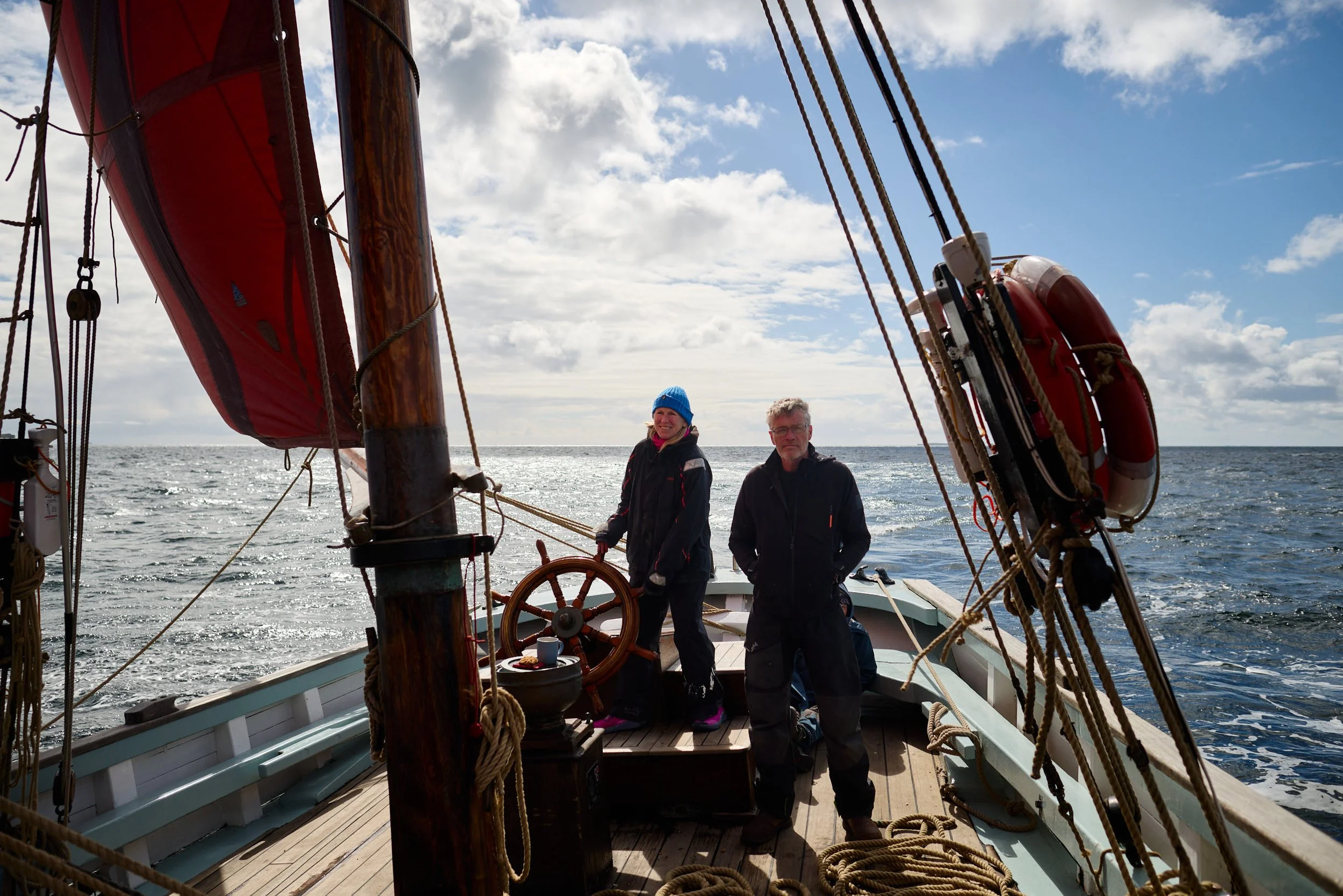 Two people standing on the deck of a sailing ship, surrounded by rigging and ropes, with the ocean and a partly cloudy sky in the background.